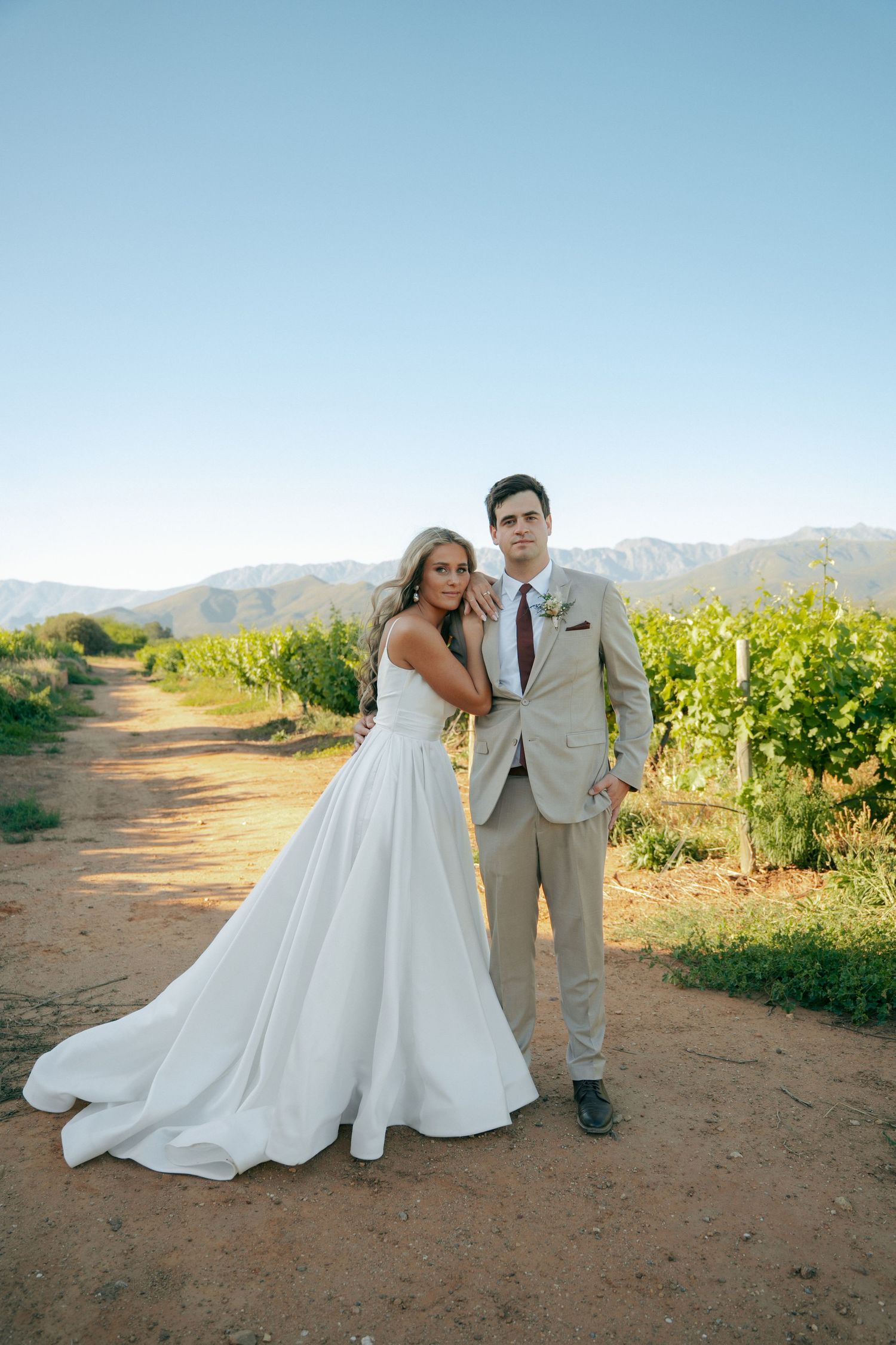 Bride in a flowing white gown leans against a groom in a tan suit on a dirt path beside green vineyards with mountains behind.