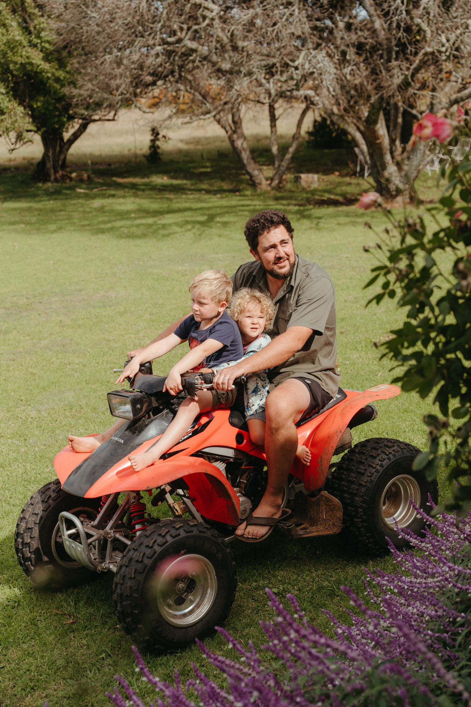 Man and two young boys riding a red ATV quad bike together on a green lawn surrounded by trees.