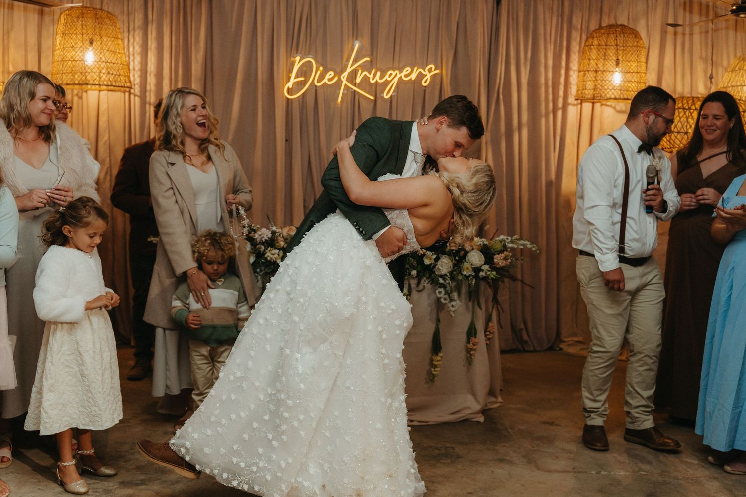 Bride and groom share romantic first dance dip kiss at wedding reception with guests watching and neon sign backdrop.