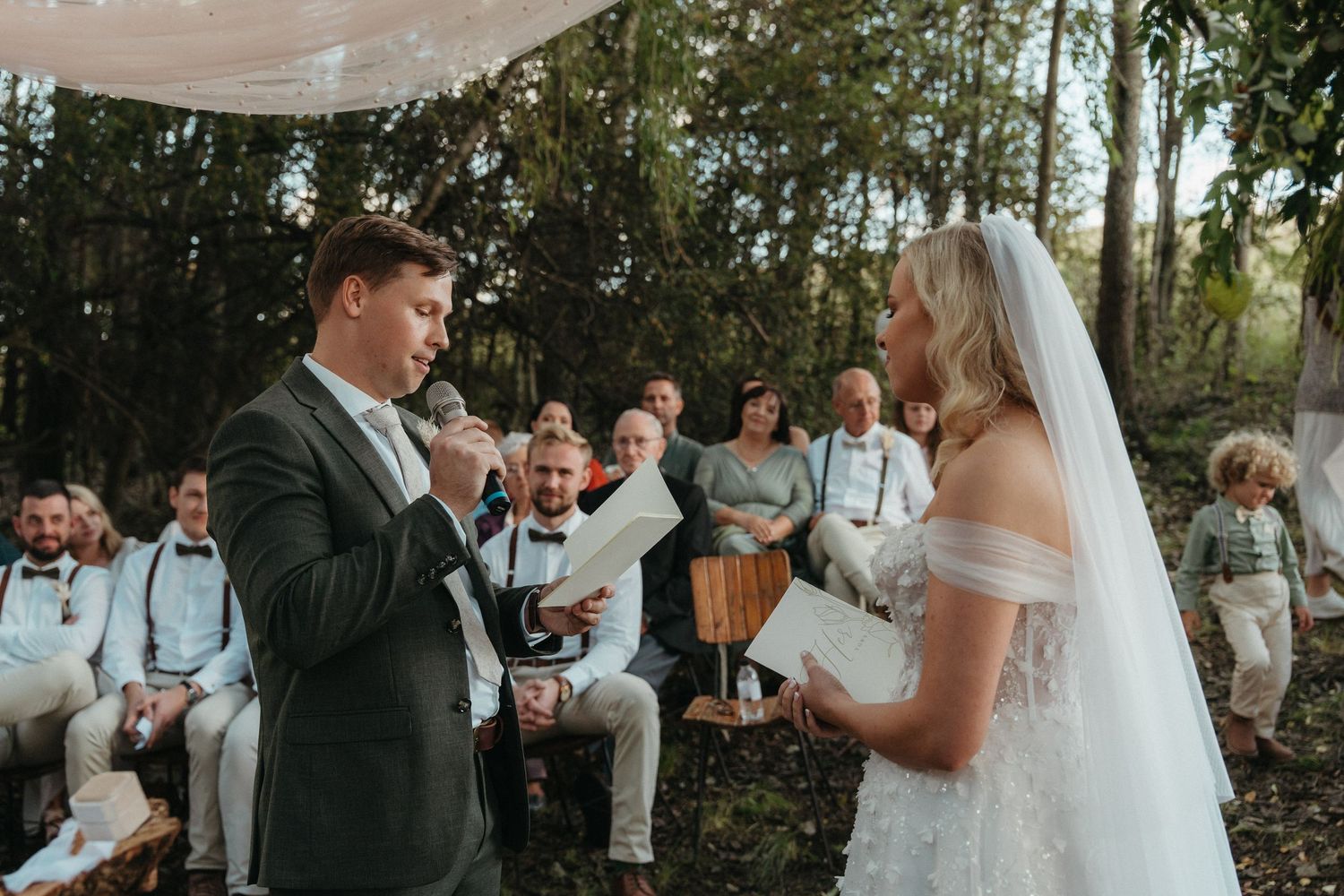 Groom reads wedding vows to bride in white dress and veil during outdoor forest ceremony with seated guests.