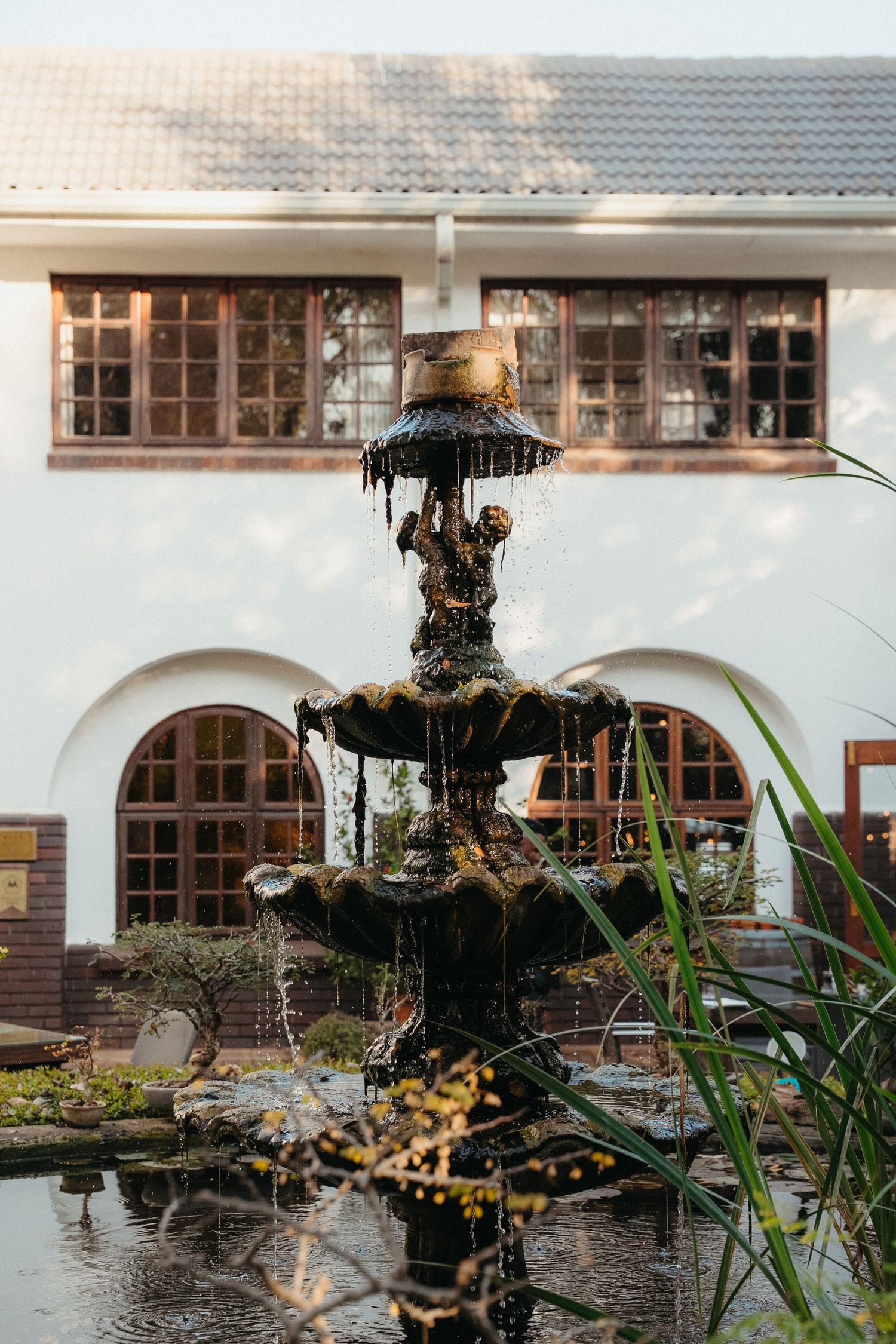 Ornate multi-tiered bronze fountain with flowing water in a lush courtyard garden beside a white building.