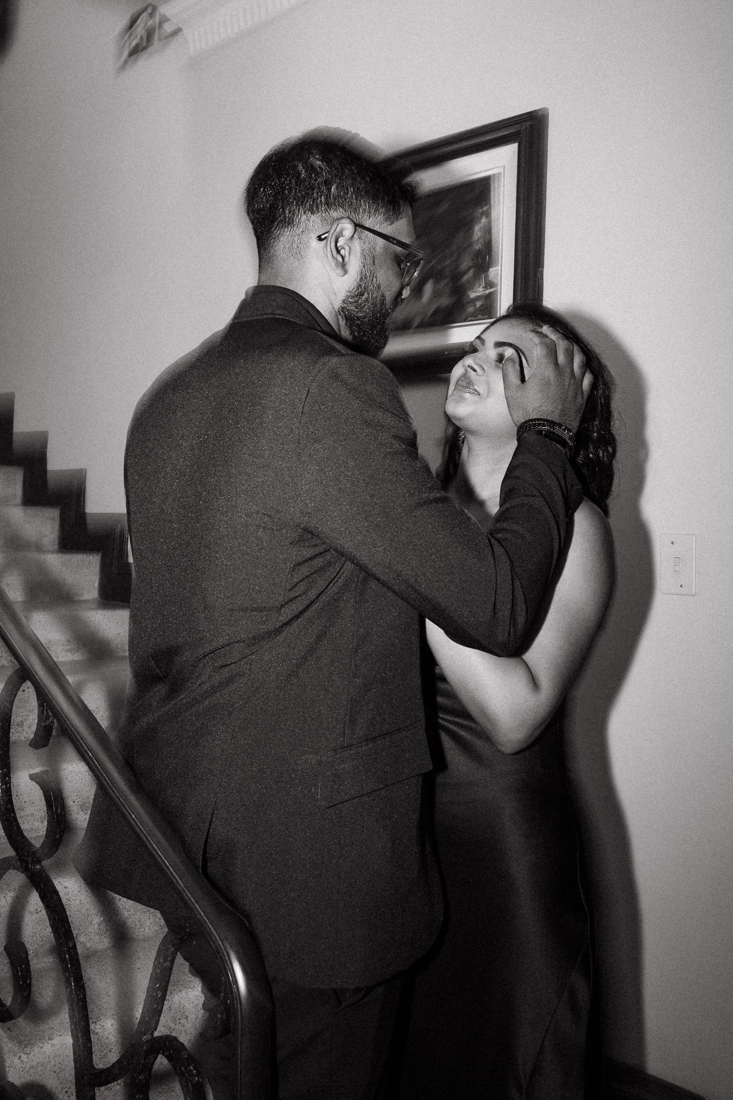 Black and white photo of a couple sharing an intimate moment near a staircase, man in suit gently holding womans face.