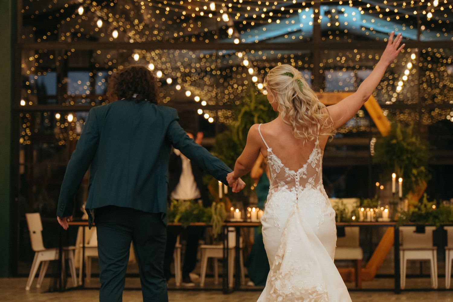 Bride in lace backless gown and groom enter Rosemary Hill's Distillery reception venue glowing with string lights and greenery in Pretoria