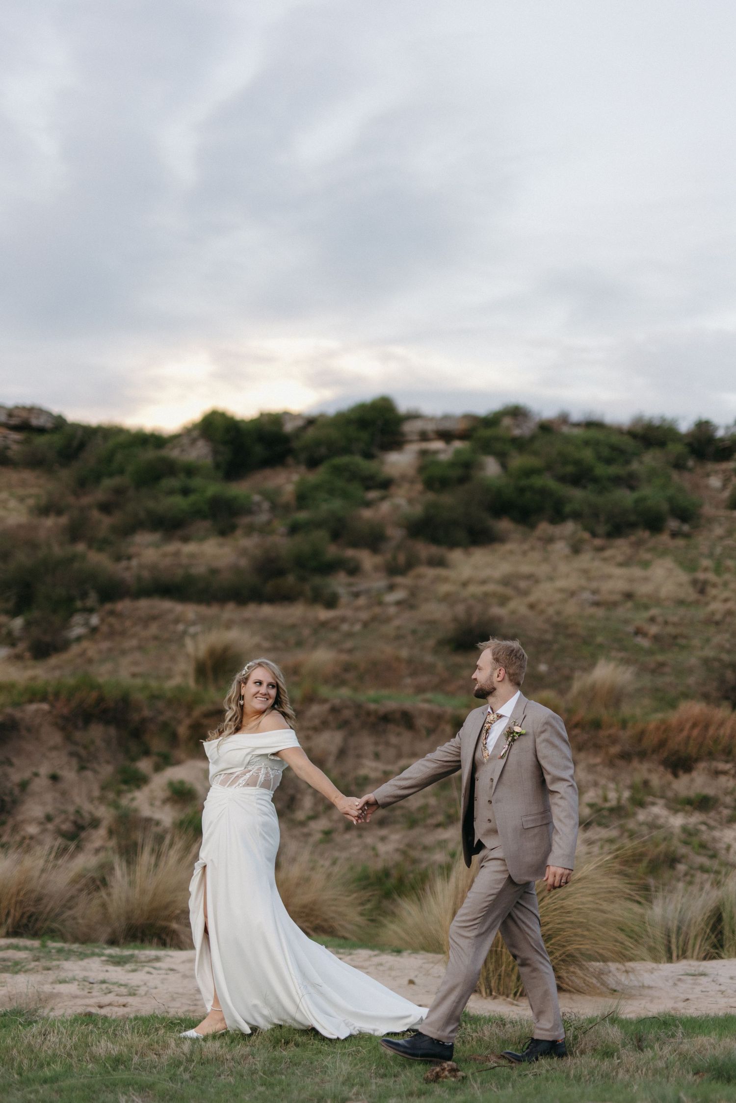 Bride and groom hold hands walking through open landscape at Smitsfield near Ermelo with dramatic cloudy sky at sunset.