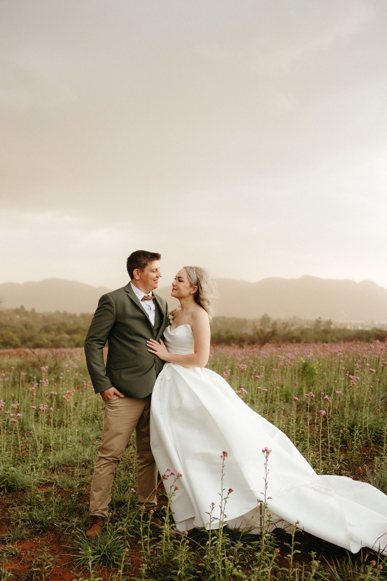 Bride and groom embrace in a blooming flower field at the Barn @ Redstone in Hartebeespoort with misty mountains in the background.