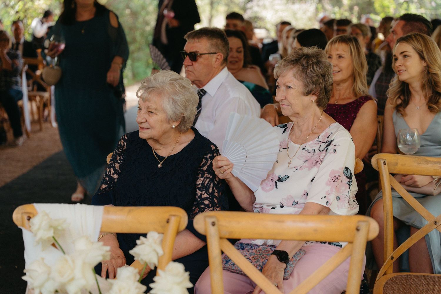 Wedding guests sit in wooden chairs during an outdoor ceremony, smiling and engaged in the celebration.
