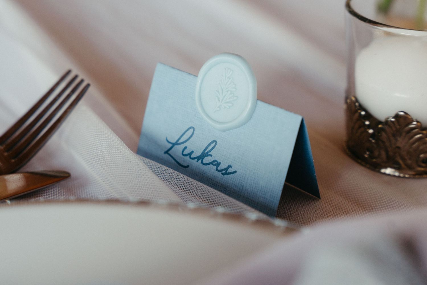 Light blue folded place card with elegant calligraphy and embossed seal on a white tablecloth setting.