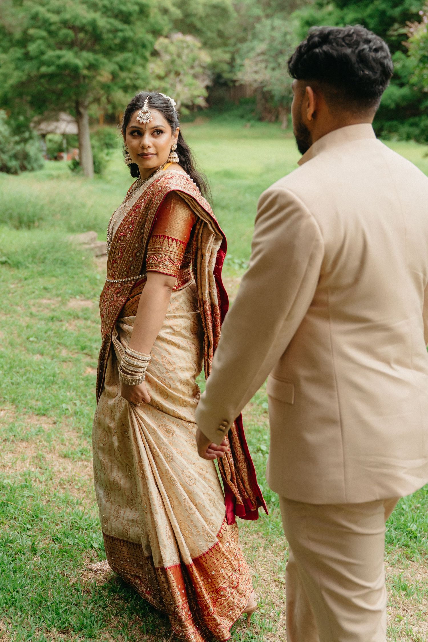 A couple in traditional South Asian wedding attire poses together in a lush green outdoor setting.