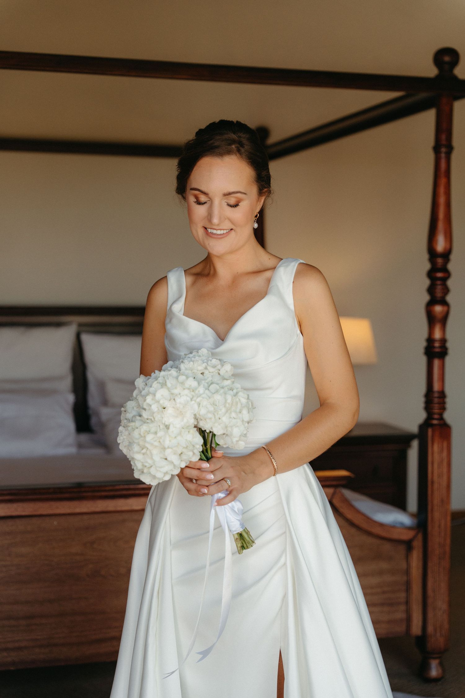 A bride in a white wedding dress holds a bouquet of white hydrangeas while getting ready in a bedroom.