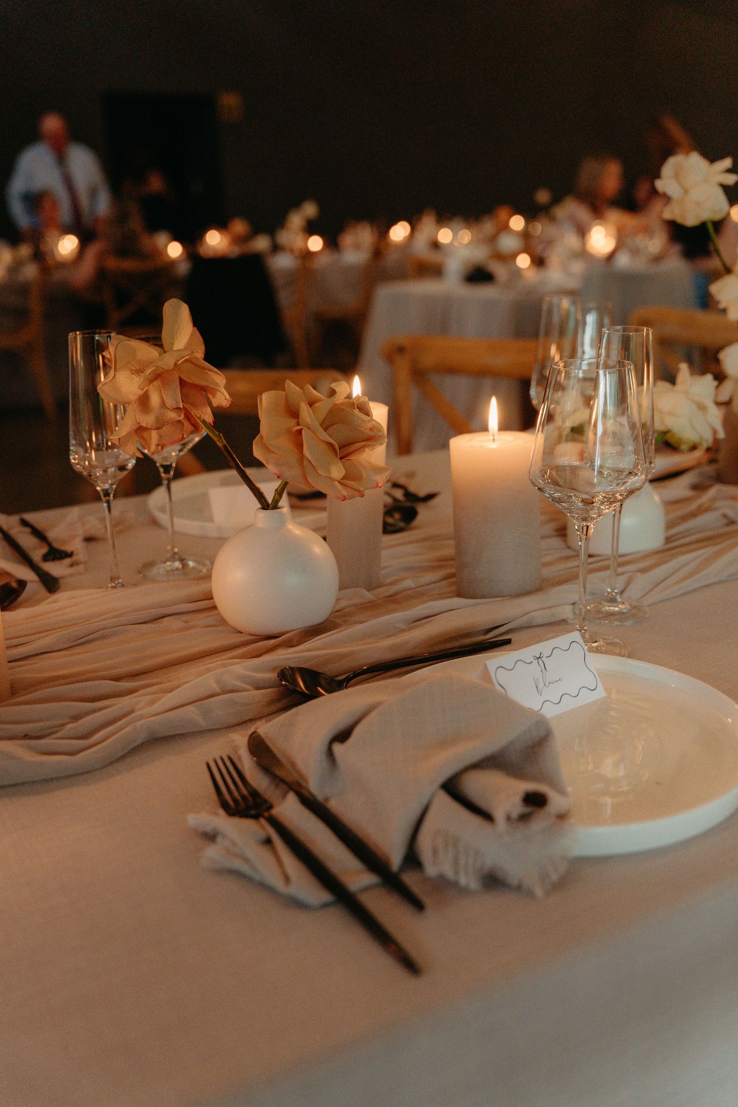 Elegant table setting with white candles, cream flowers, and place settings on a white tablecloth in warm lighting.