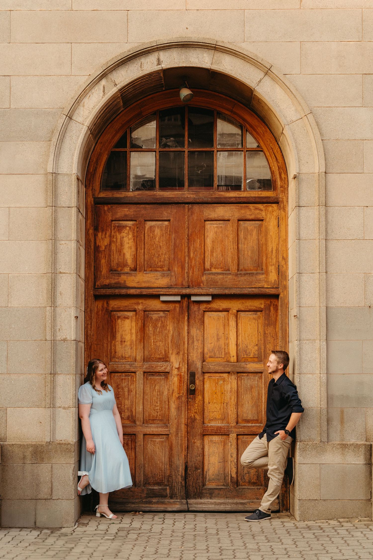 Couple poses against large vintage wooden arched door on stone building in warm evening light.