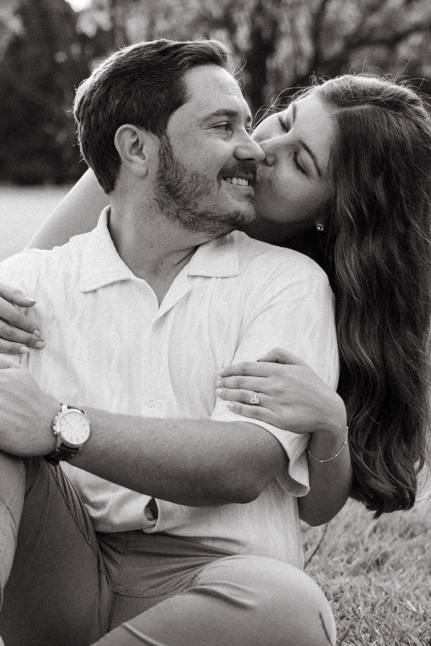 Black and white photo of a smiling man wearing a watch as a woman with dark hair kisses his cheek while embracing him.