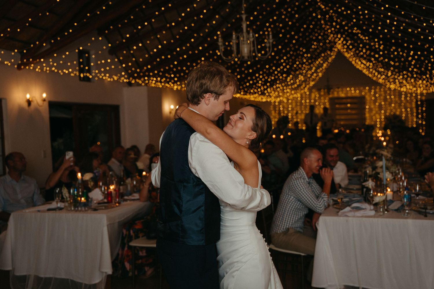 A couple shares a romantic dance under twinkling string lights at an indoor wedding reception with tables in the background.