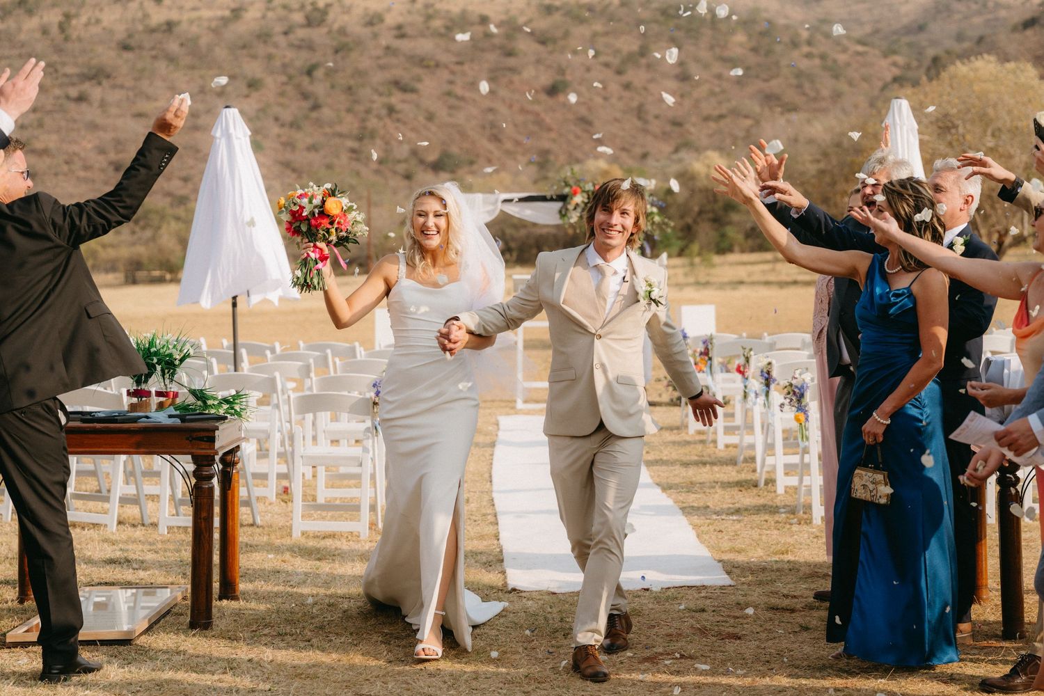 Bride and groom walk down outdoor aisle as guests raise hands in celebration under sunny Limpopo sky at Mountain View Game Lodge