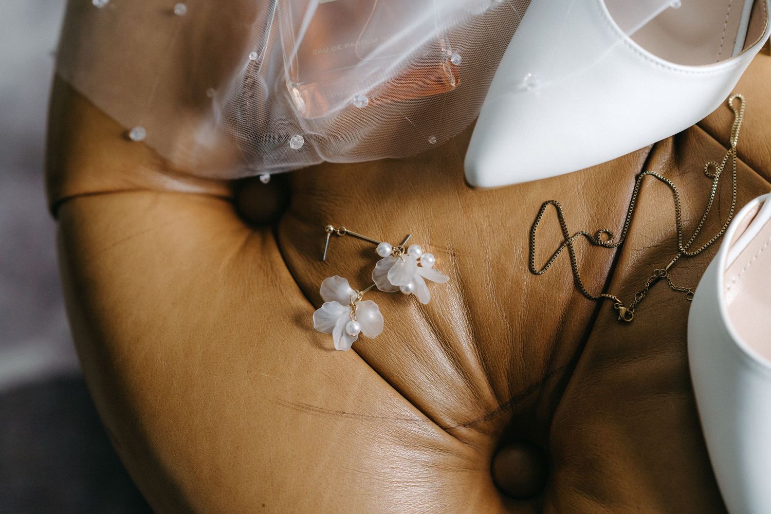 White bridal shoes and pearl earrings arranged on brown leather surface.