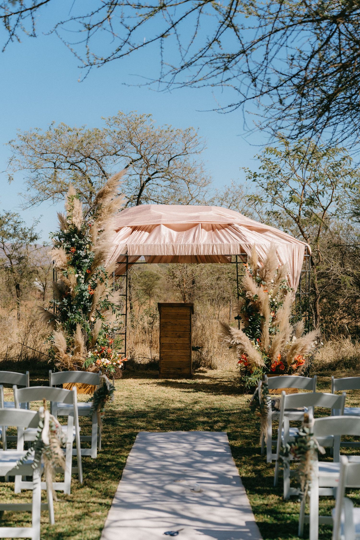 Outdoor wedding ceremony setup with white chairs and a blush pink canopy against a natural wooded backdrop.