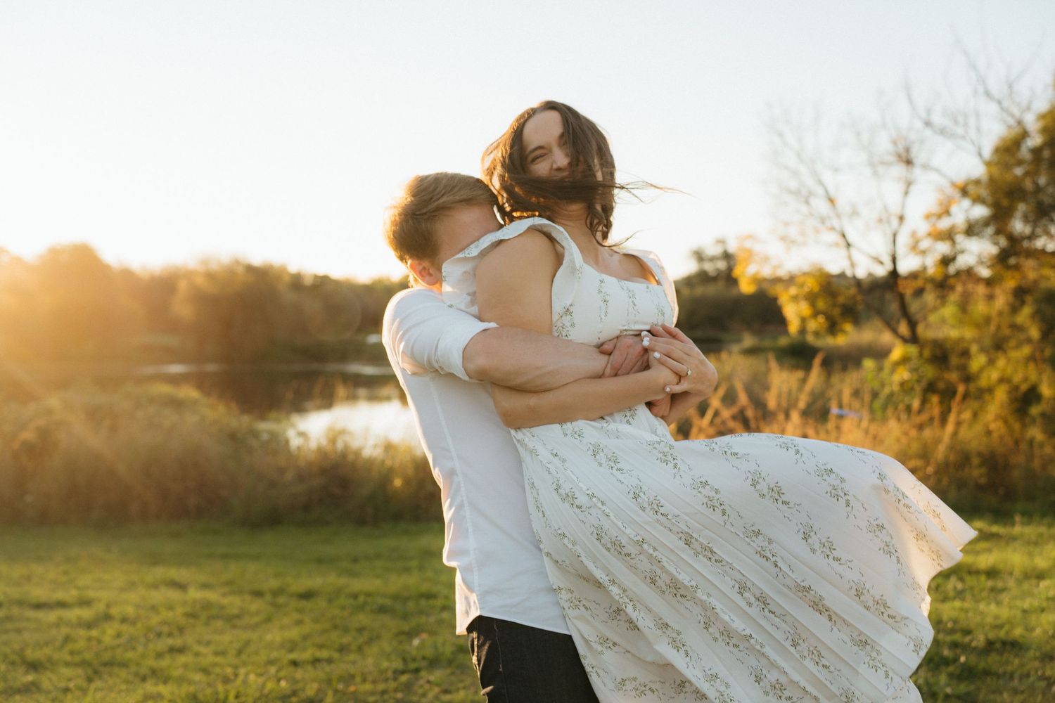 A couple embraces in a sunlit field during golden hour as her white lace dress flows in the breeze.