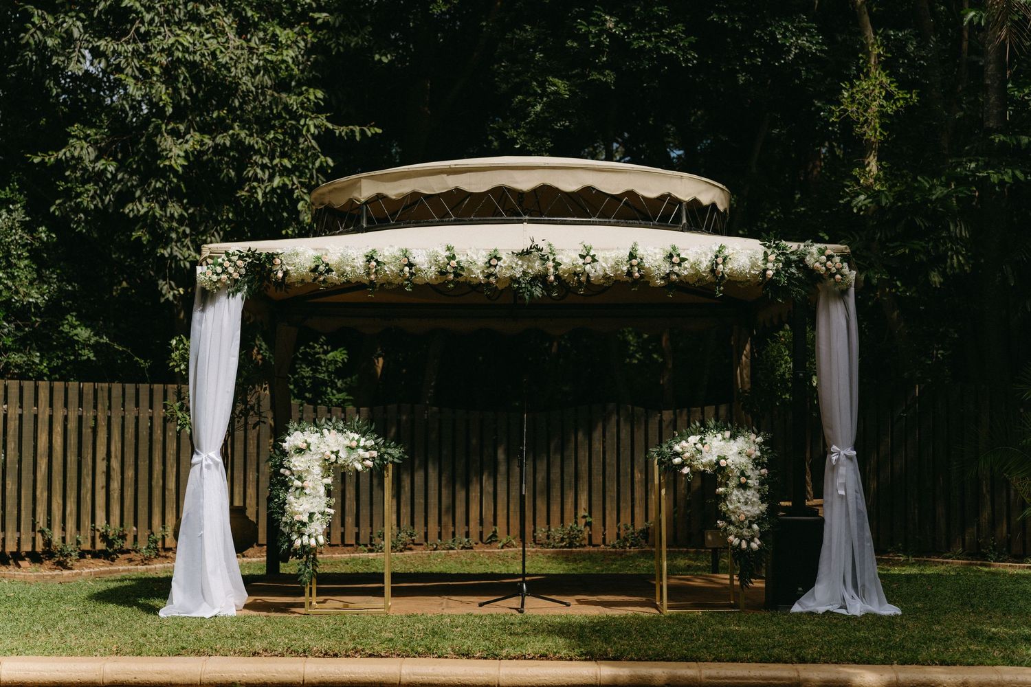 A rustic wooden wedding arch decorated with white fabric draping and floral arrangements stands in an outdoor garden setting.