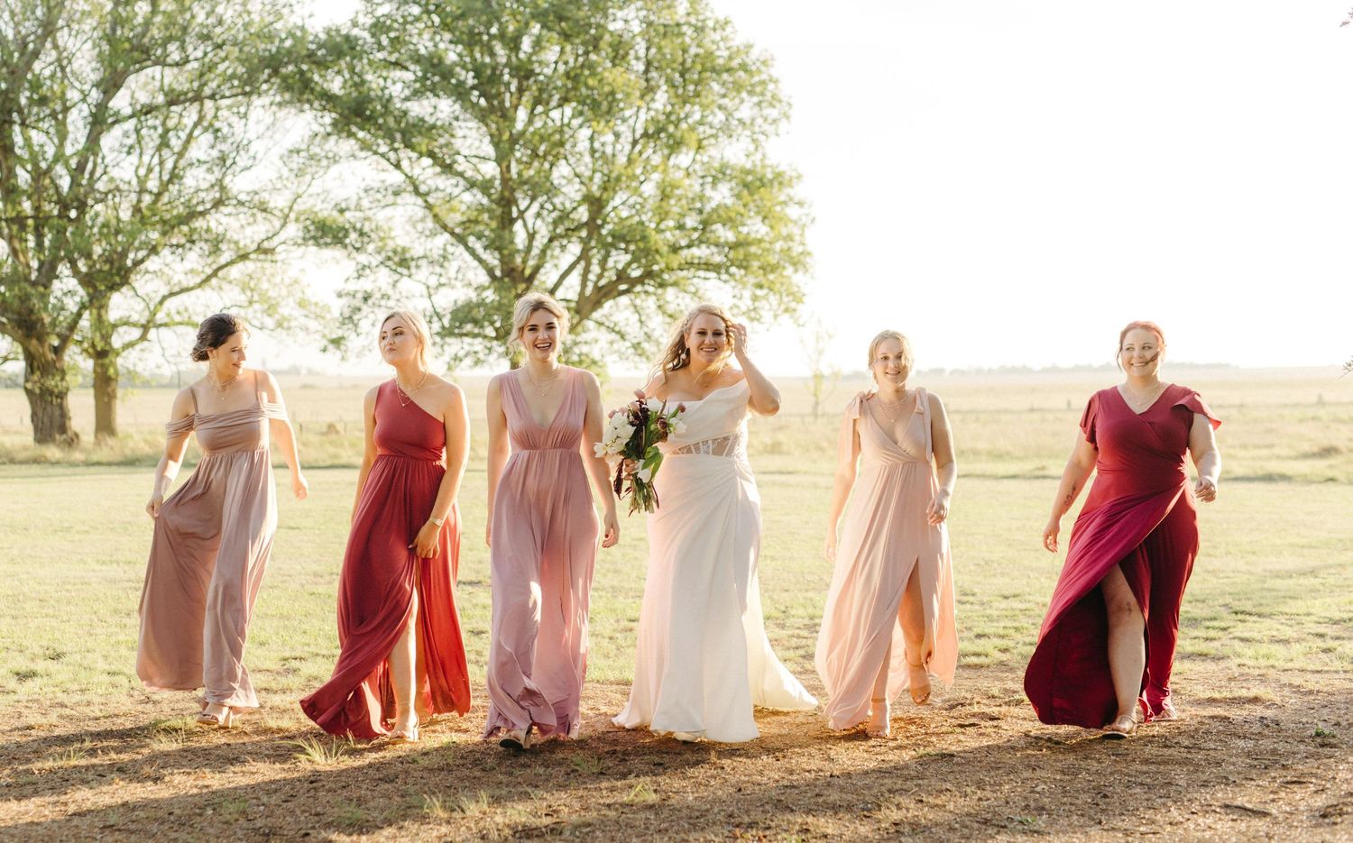 Group in coordinated pink and red formal dresses pose together in a sunlit field.