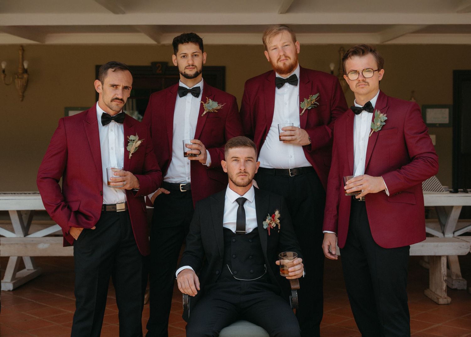 Groomsmen in burgundy tuxedo jackets and bow ties posing together in an elegant indoor setting.