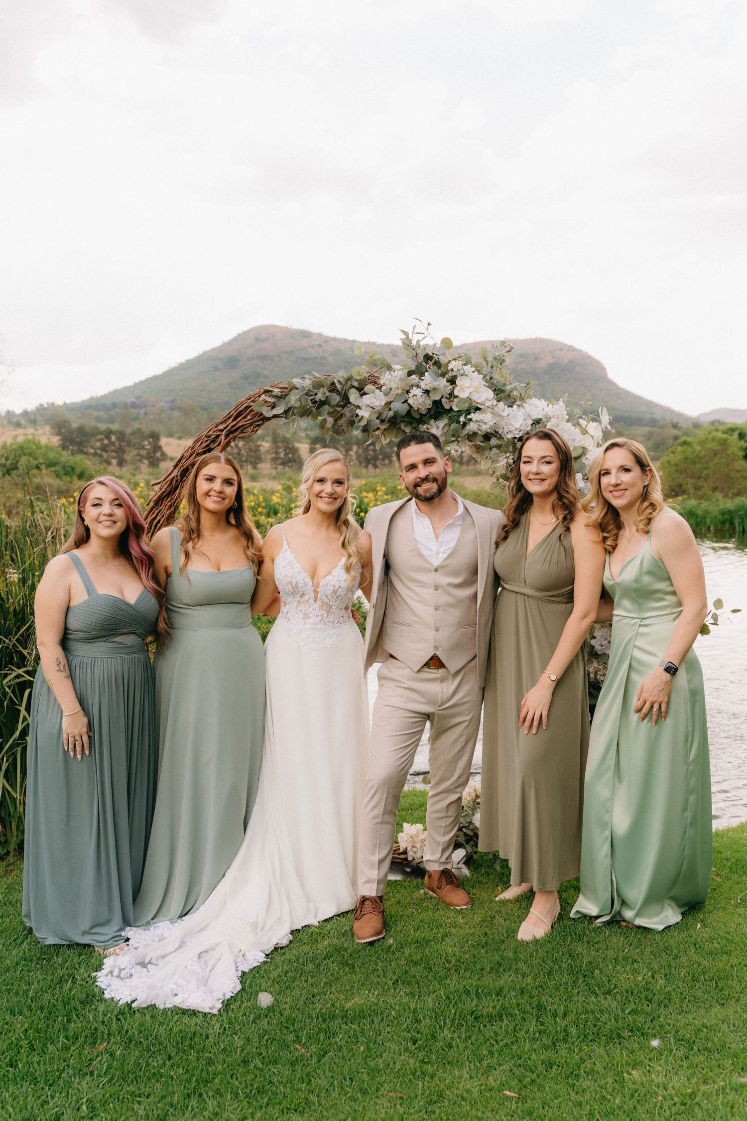 Wedding party poses outdoors in sage green and neutral dresses against mountain backdrop.