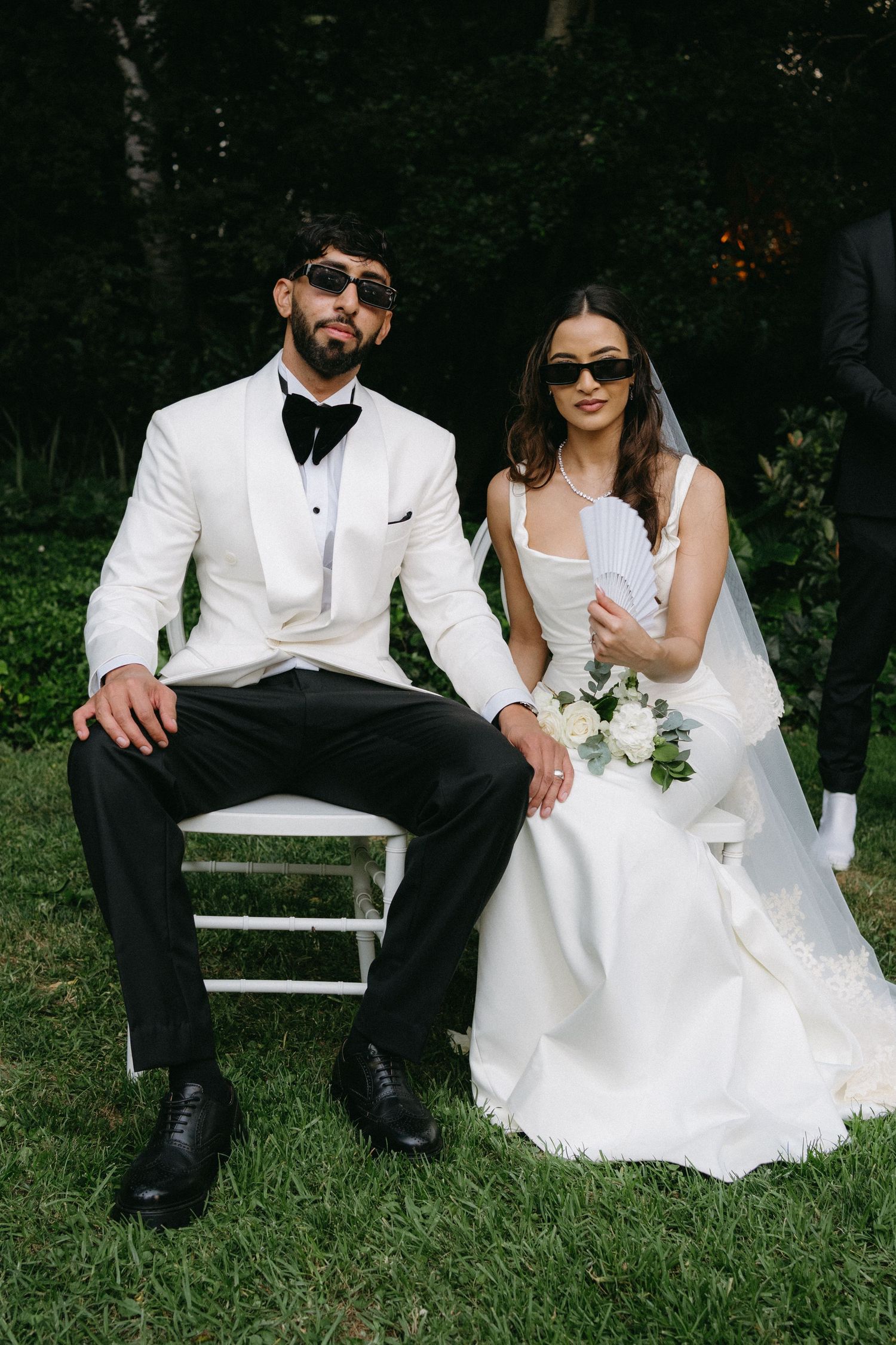 Newlyweds in white attire pose stylishly on chairs outdoors, both wearing sunglasses with a bouquet nearby.