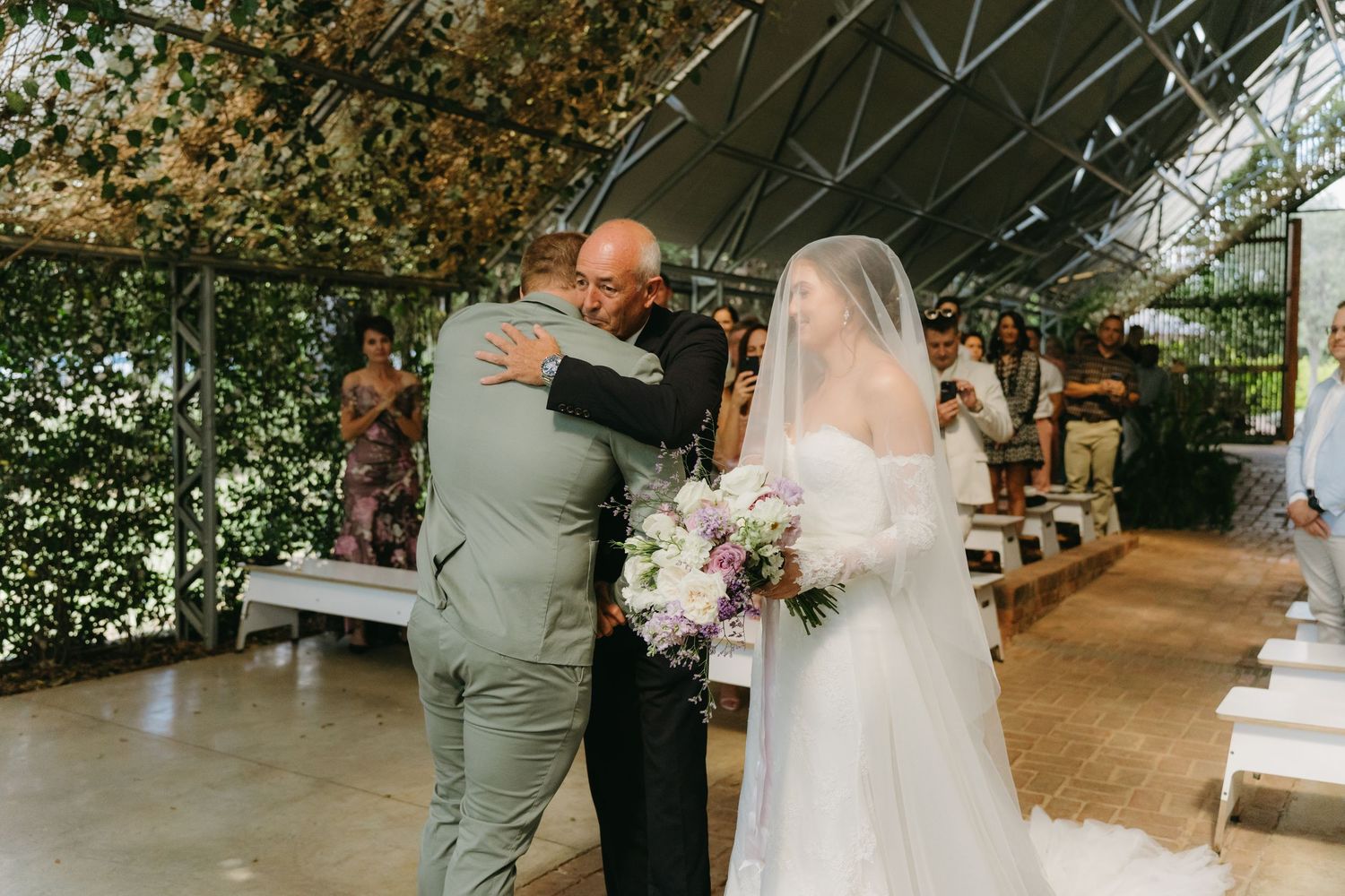 A wedding ceremony takes place under a covered outdoor venue with guests seated on white benches.