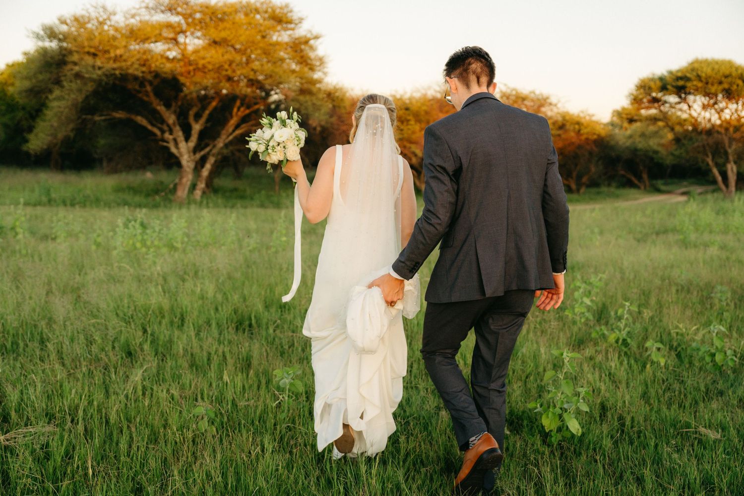 A newlywed couple walks through a grassy field at sunset with golden autumn trees in the background.