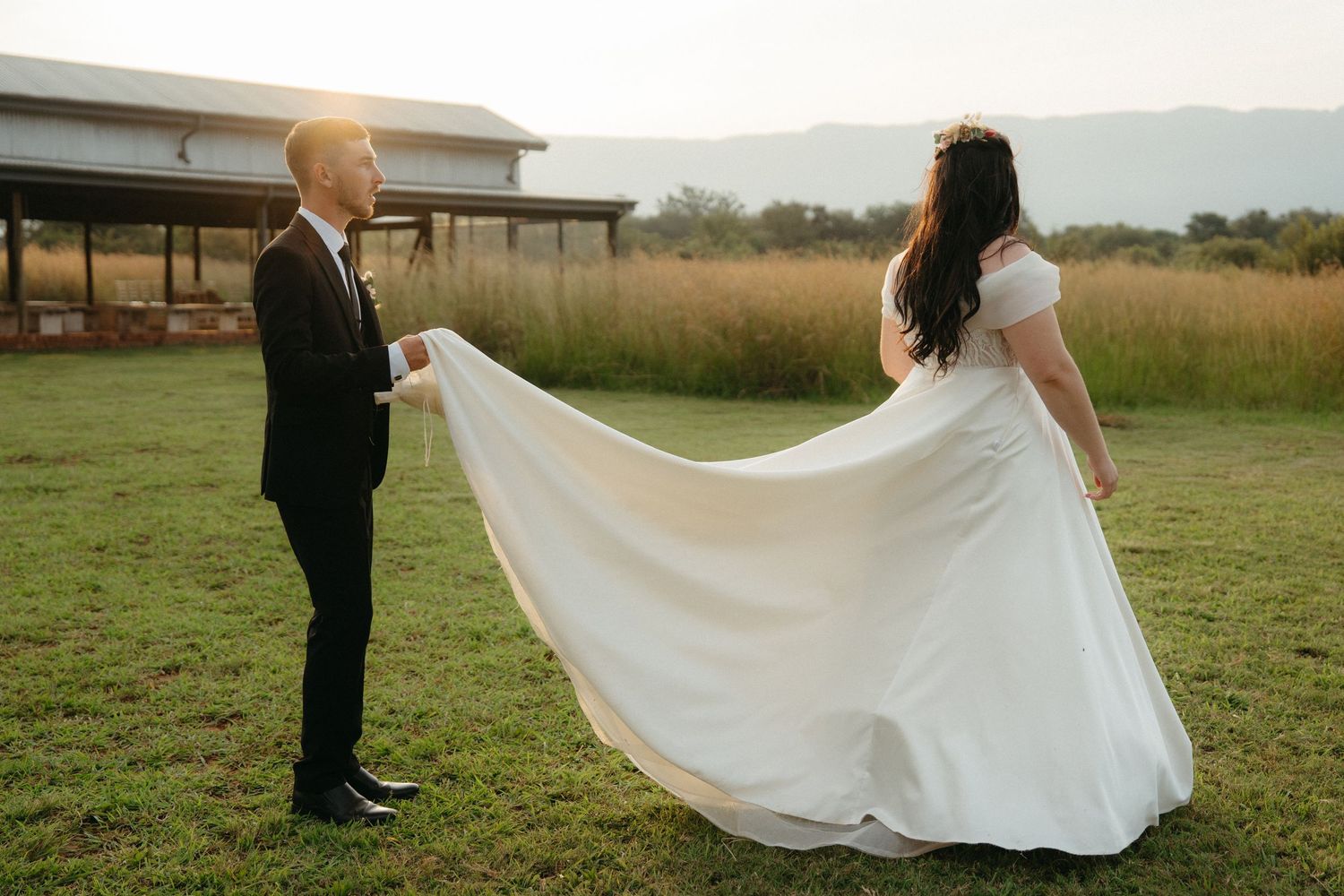 A wedding couple stands in a grassy field as the groom helps adjust the bride's flowing white dress.