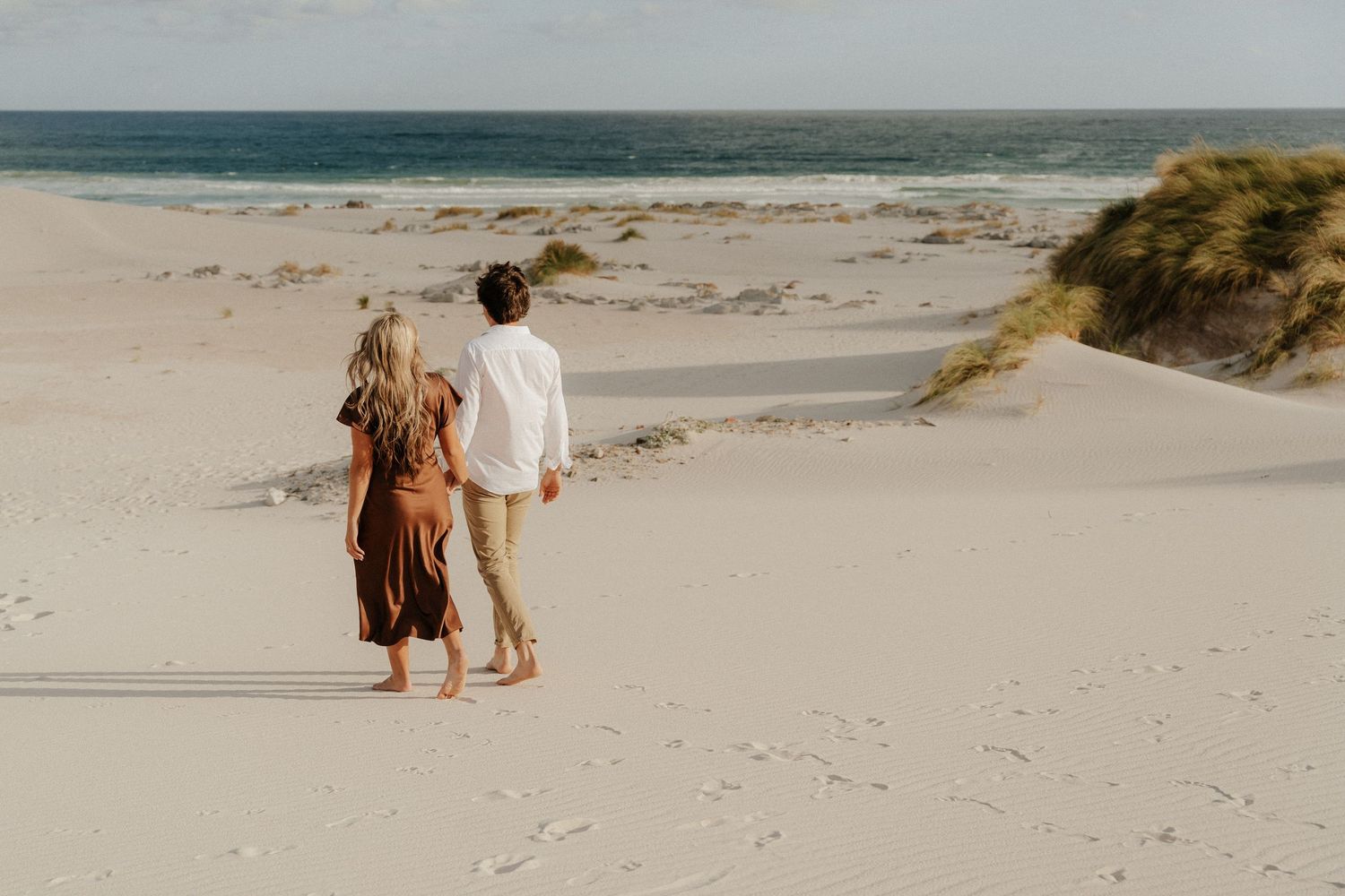 Couple holding hands walking barefoot on white sand dunes near the ocean with green grass in background.