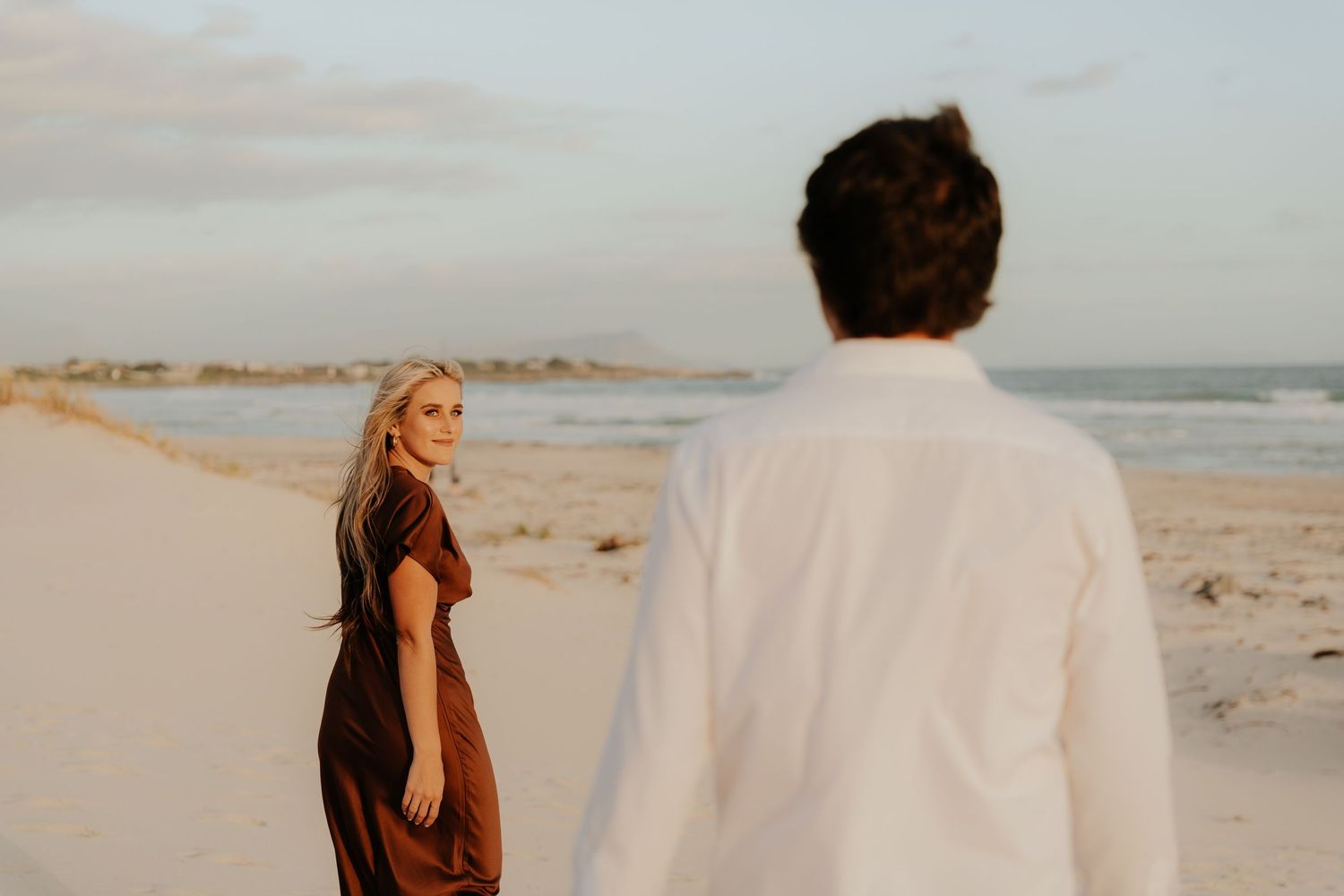 A blonde woman in a brown dress looks back at a man in a white shirt on a sandy beach at golden hour.