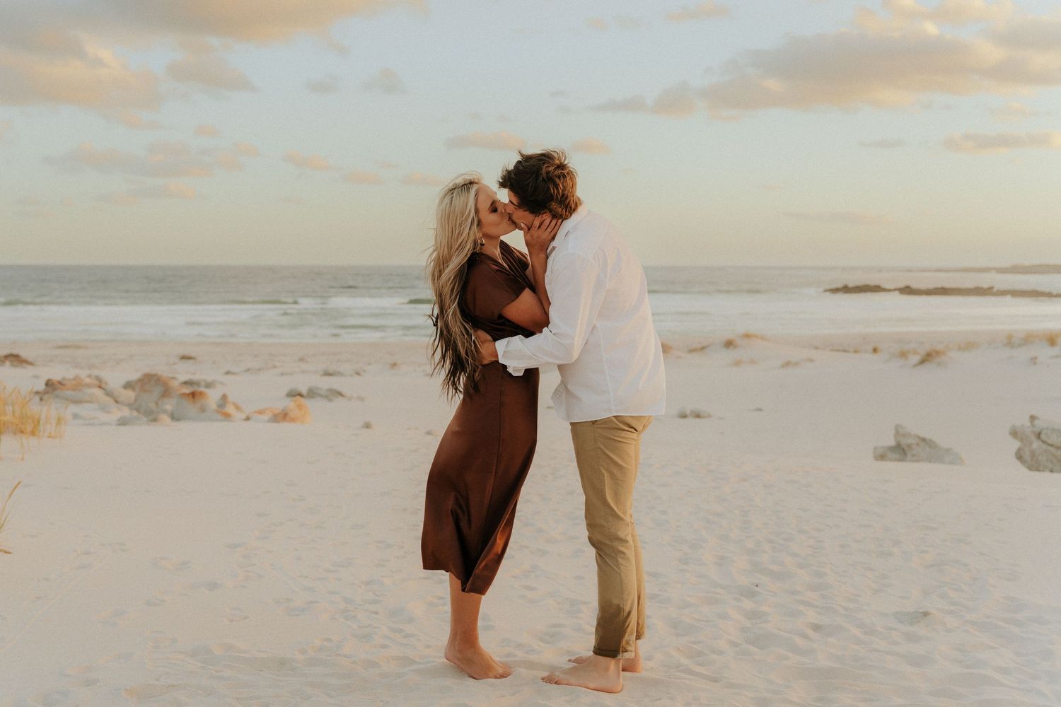 A couple shares a romantic moment on a sandy beach at sunset, the woman in a brown dress and man in white.