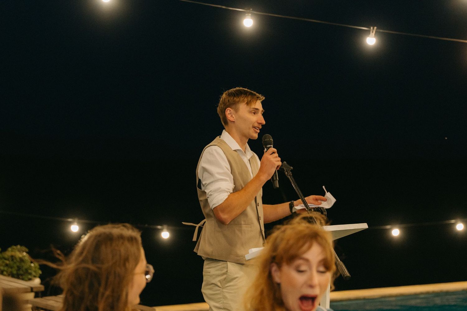 Evening wedding reception under string lights with guests seated at tables outdoors.