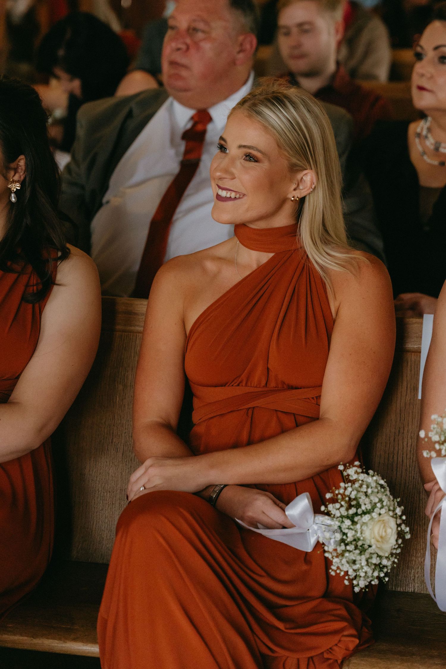 Wedding attendants in rust-colored dresses hold delicate baby's breath bouquets during ceremony.