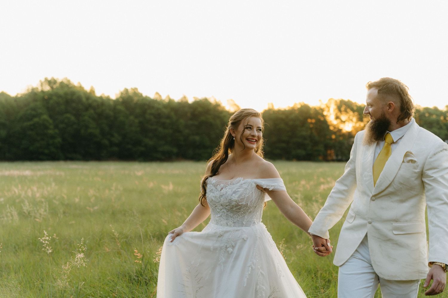 Newlyweds walk hand in hand through a sunlit field during golden hour.