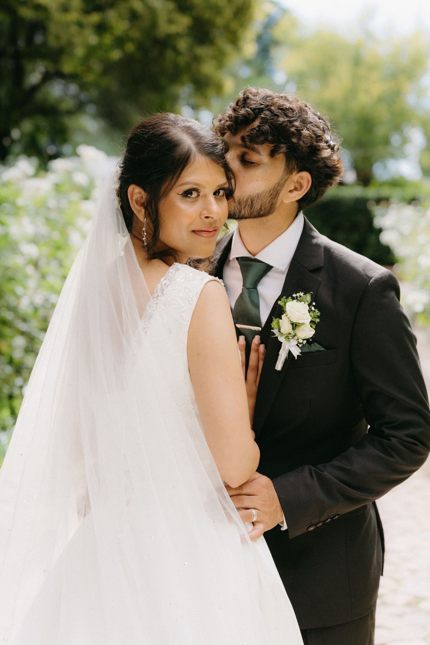 A couple in wedding attire share an intimate moment in a sunlit garden setting.