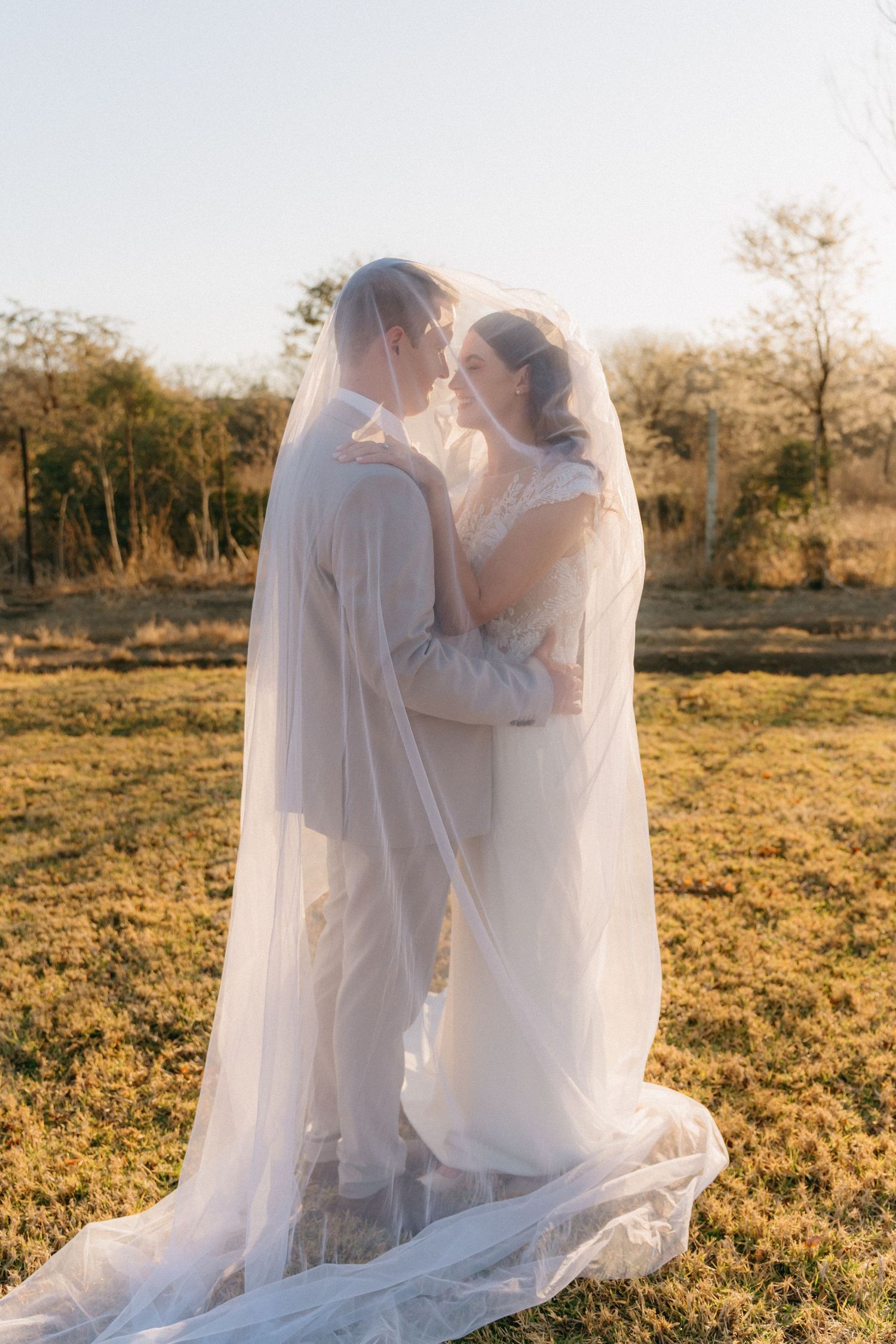 A romantic wedding couple embraces in a sunlit field with the bride's veil flowing behind them.
