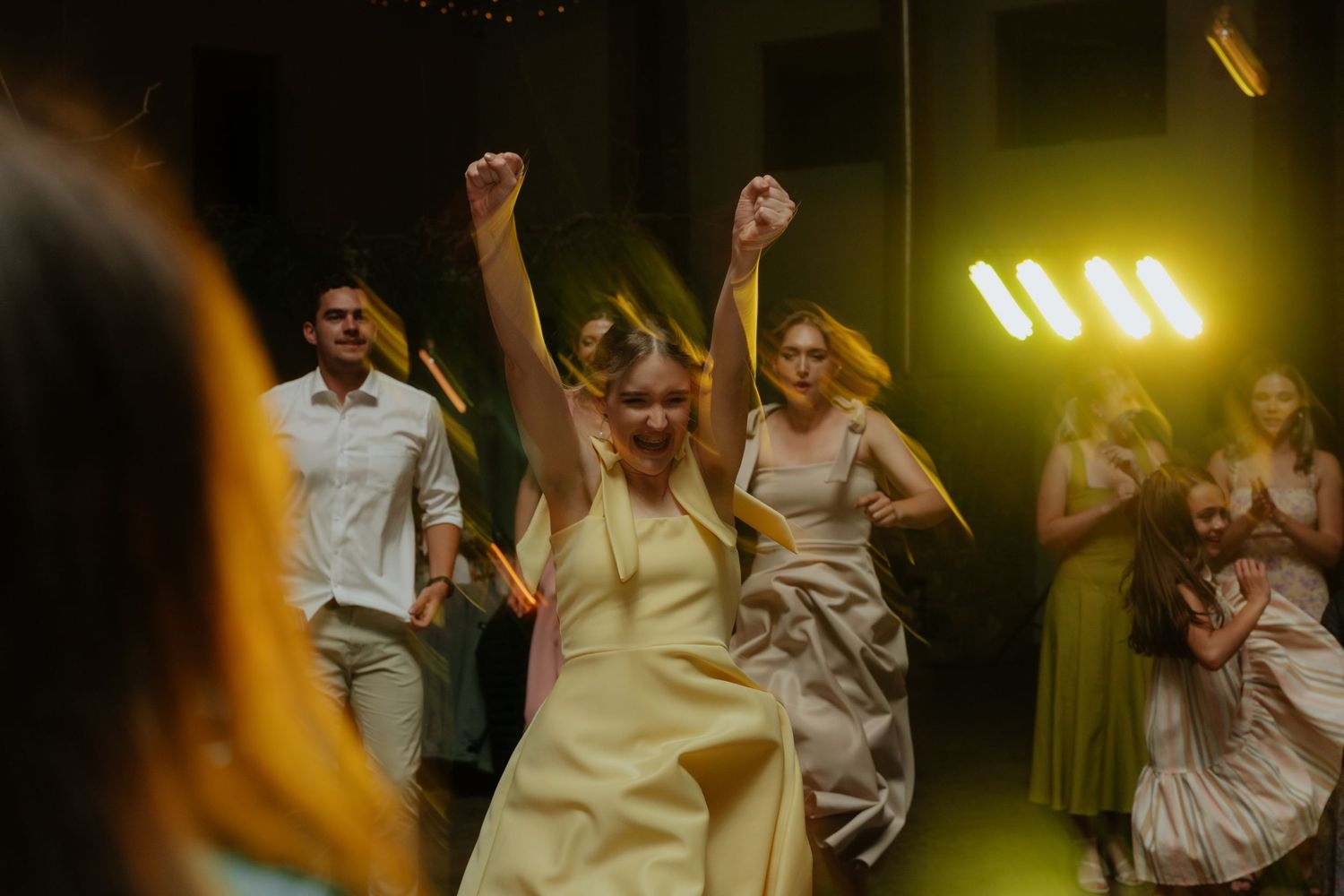 Wedding guests dance and celebrate with raised arms on a dimly lit dance floor during a reception party.