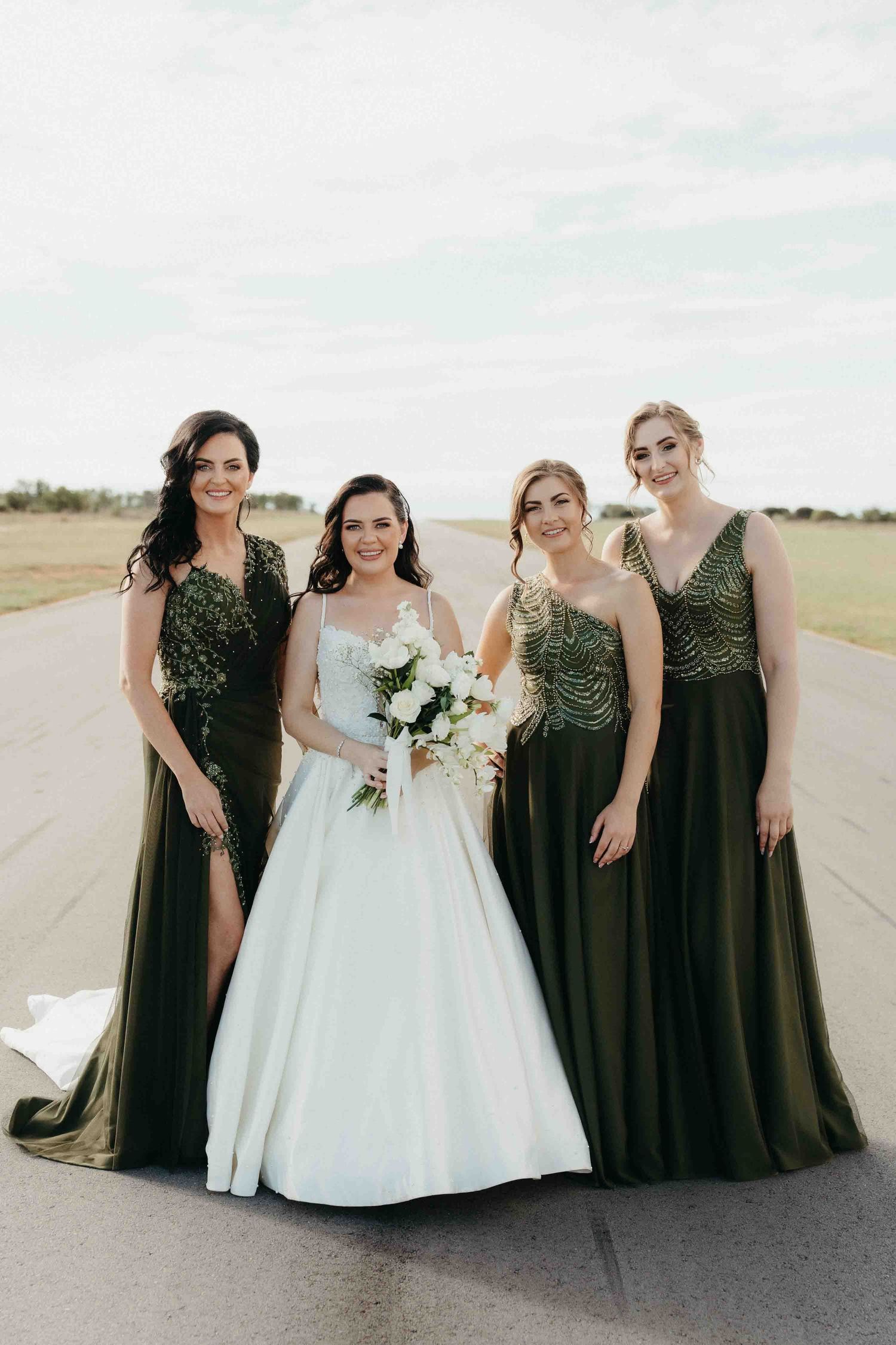 Bride in white gown holds bouquet with three bridesmaids in olive green embellished dresses outdoors.