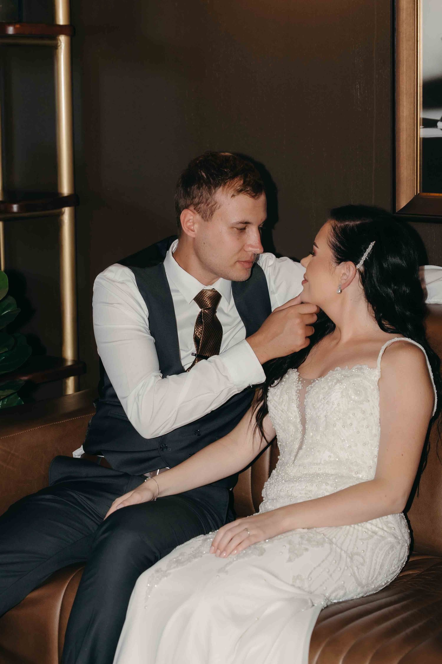 Groom in navy vest gently touches bride's face while sitting together on a leather sofa in an elegant venue.