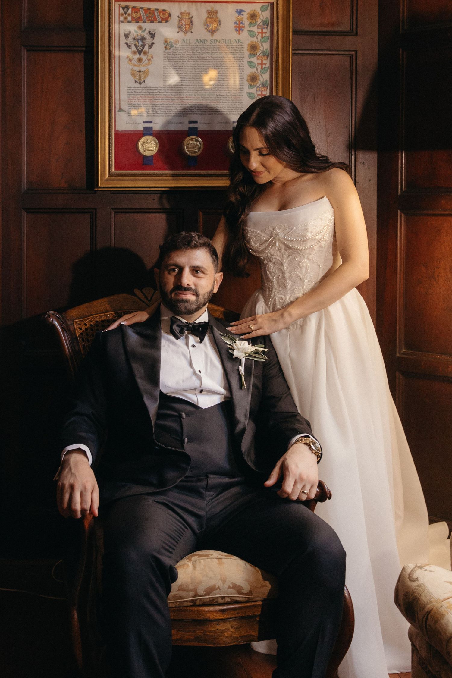 Bride in strapless white gown stands behind groom in black tuxedo seated in vintage chair in elegant wood-paneled room.