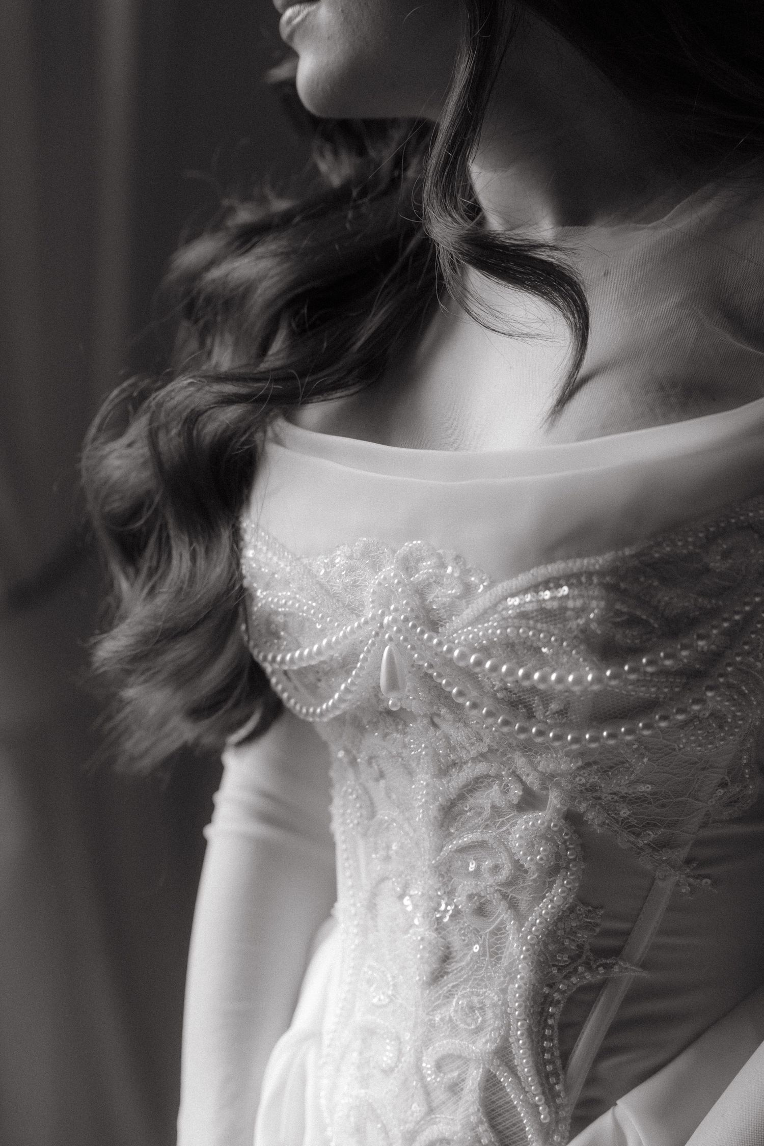 Black and white photo of a bride in an elegant beaded lace wedding gown with off-shoulder neckline and curled hair.