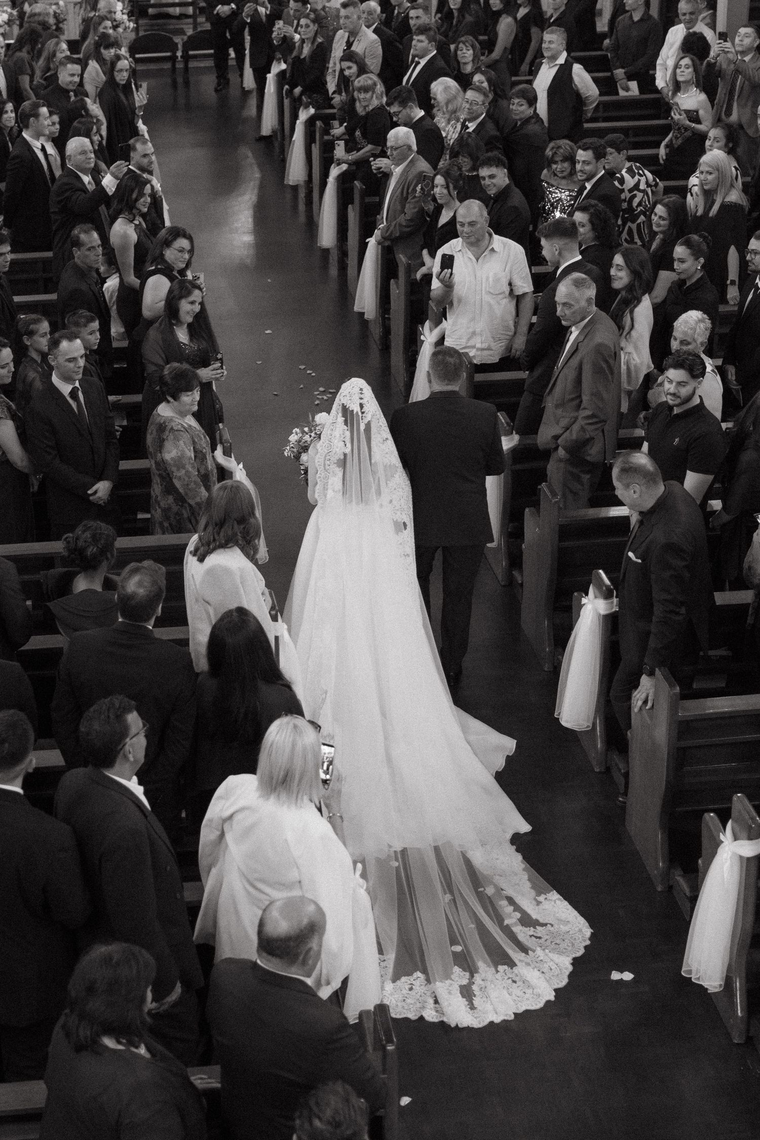 Black and white photo of a bride walking down the Rosebank Catholic Church aisle in a long veil, surrounded by wedding guests.