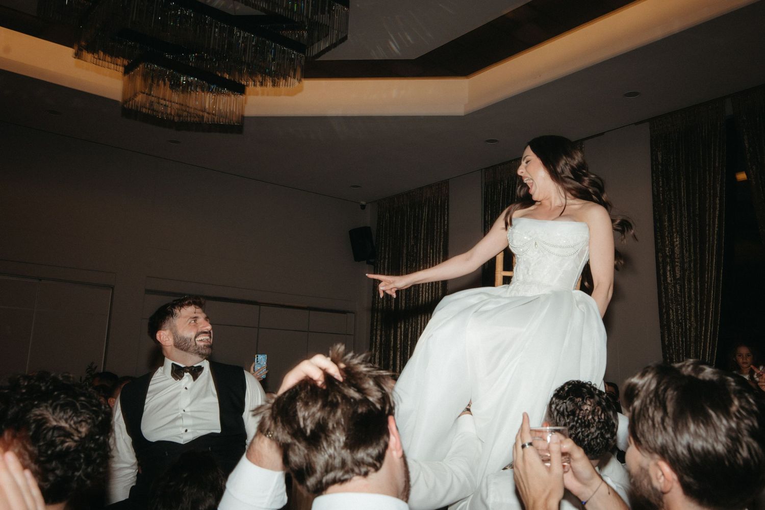 Bride lifted by guests on wedding dance floor while groom laughs joyfully during lively wedding reception celebration.