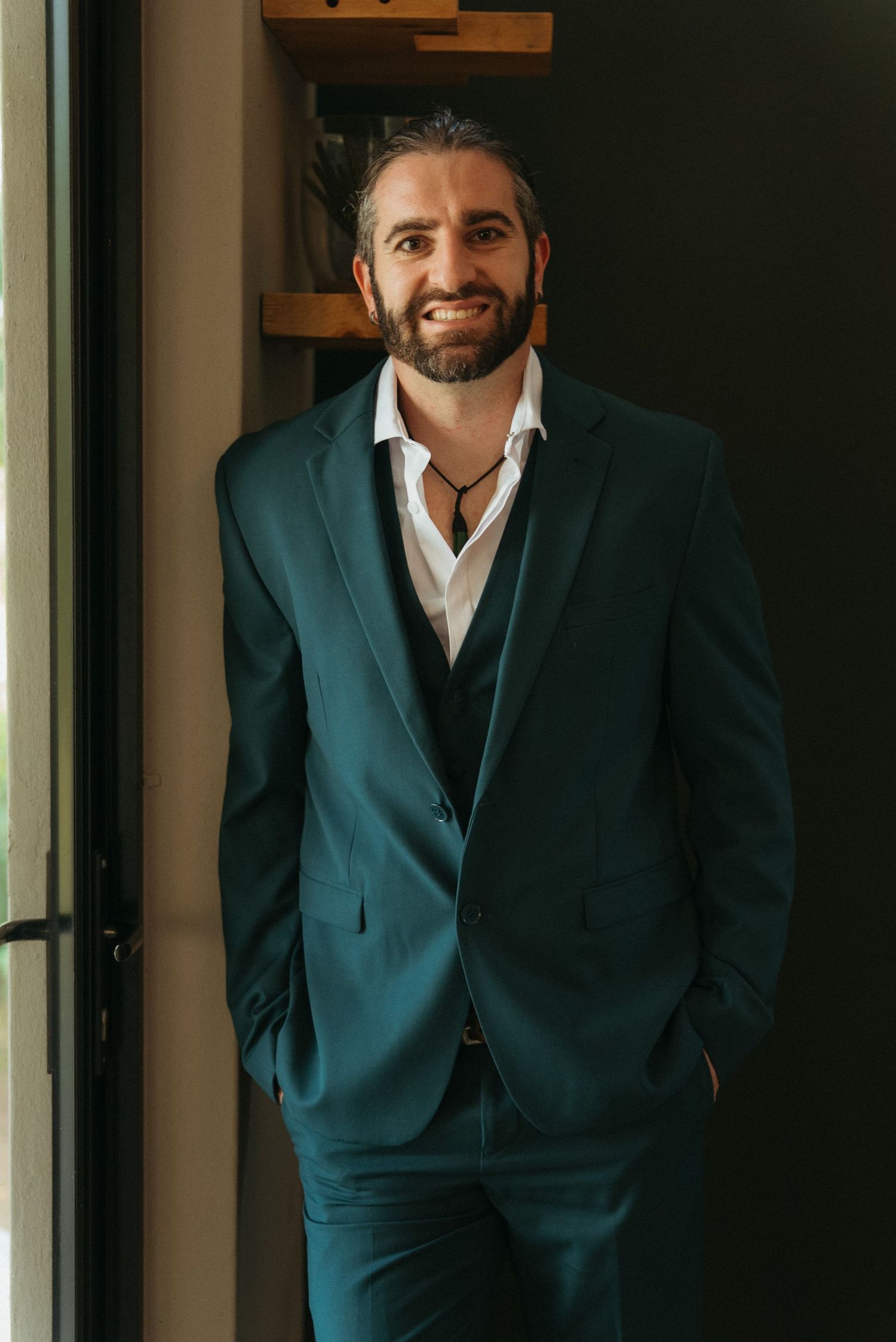 Smiling bearded man in a teal three-piece suit with white shirt, posing confidently indoors.