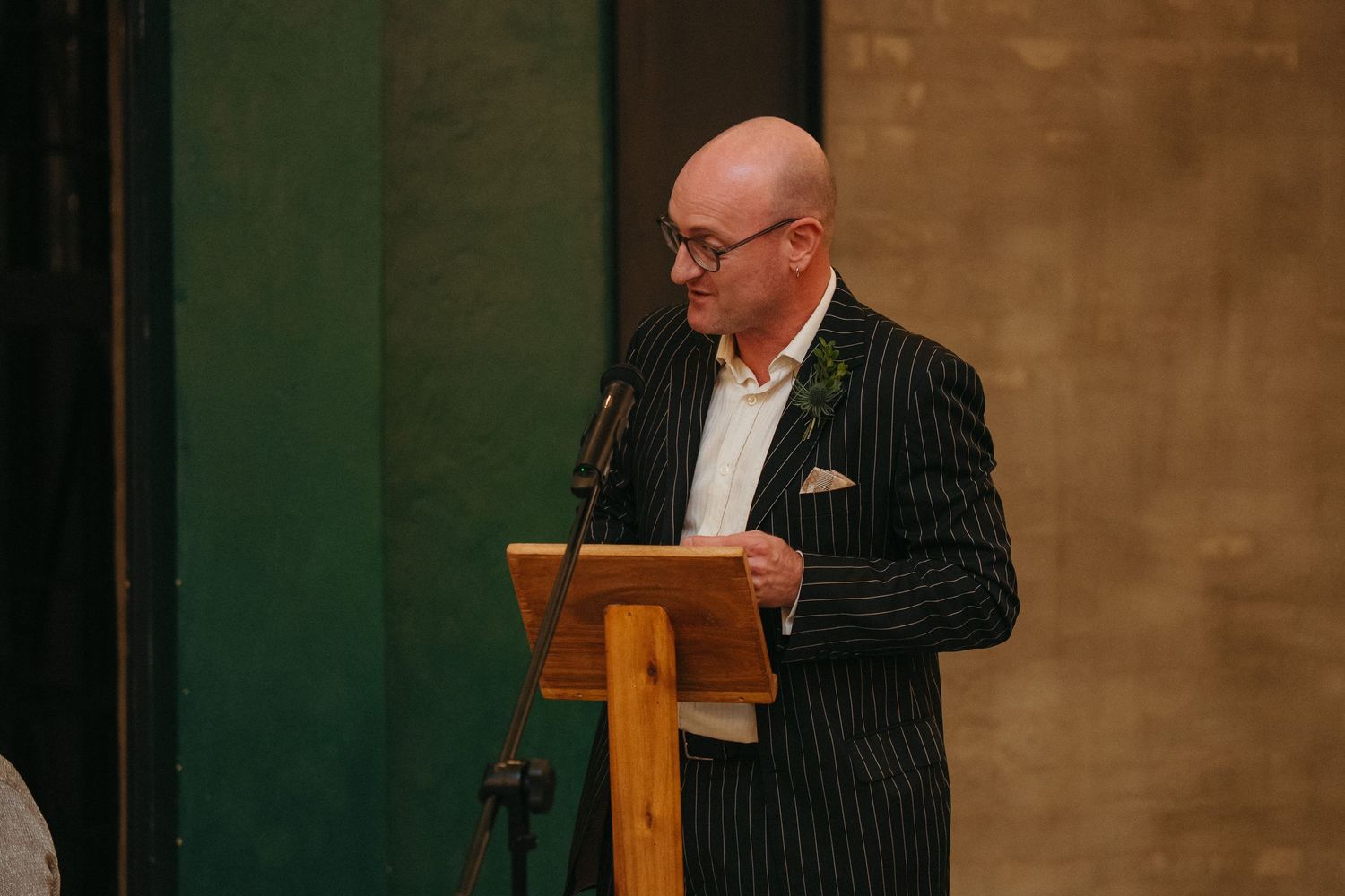 Bald man in pinstripe suit speaking at wooden podium with microphone at formal event.