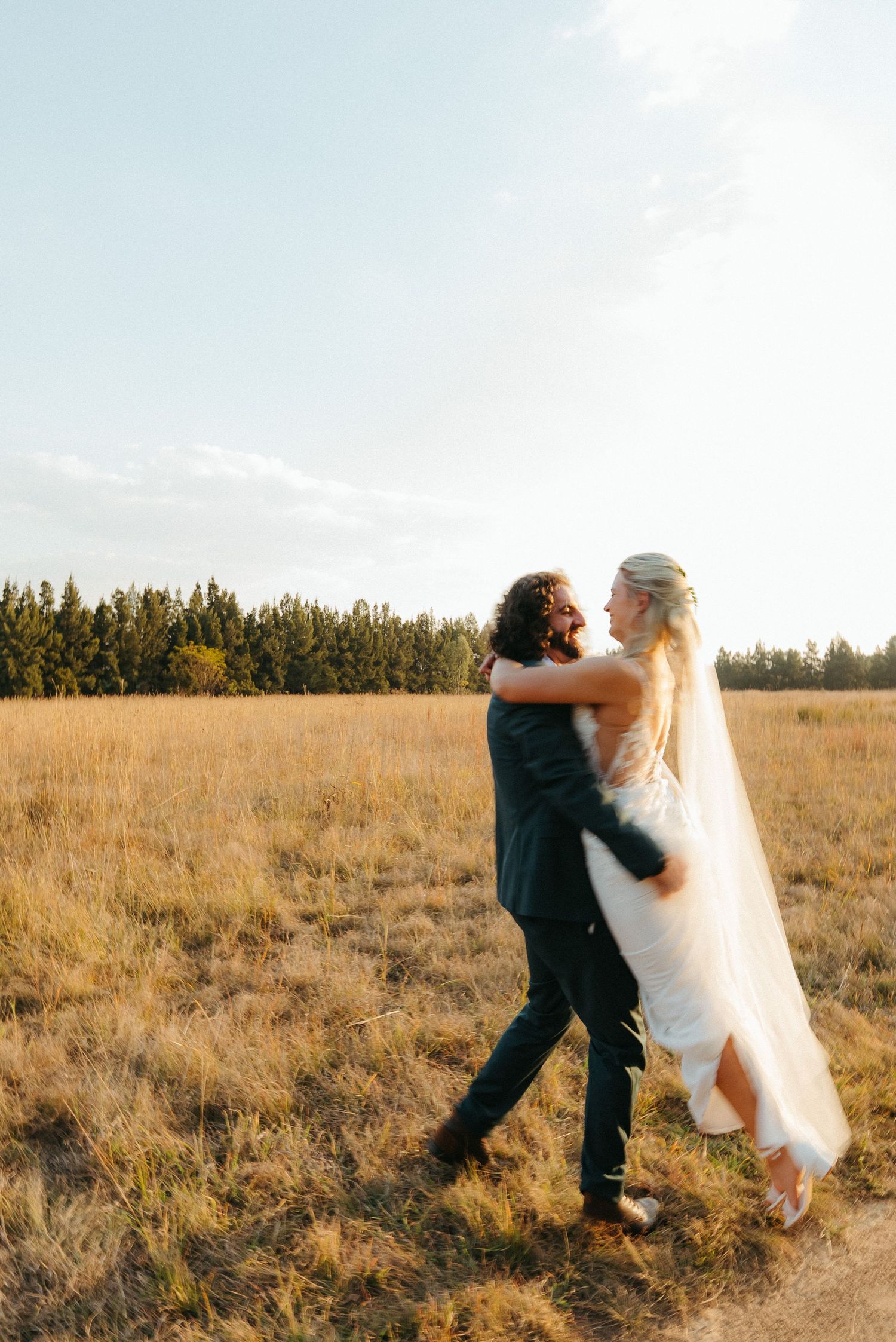Bride and groom embracing in golden field at sunset, wearing wedding dress and dark suit, with pine trees in background.