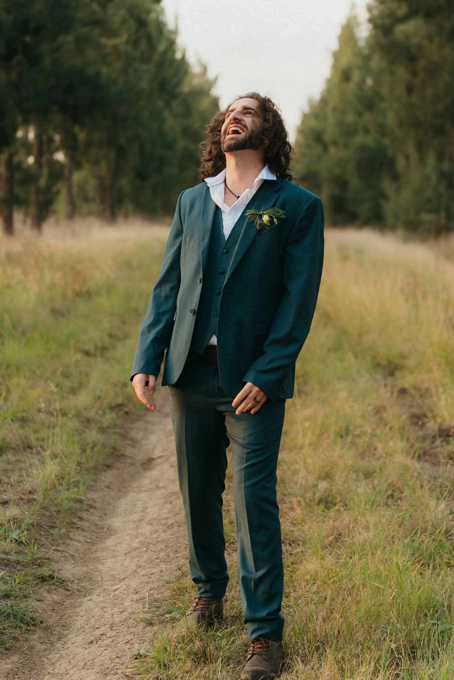 Laughing groom in green suit with boutonniere walking on dirt path through forest field on wedding day.