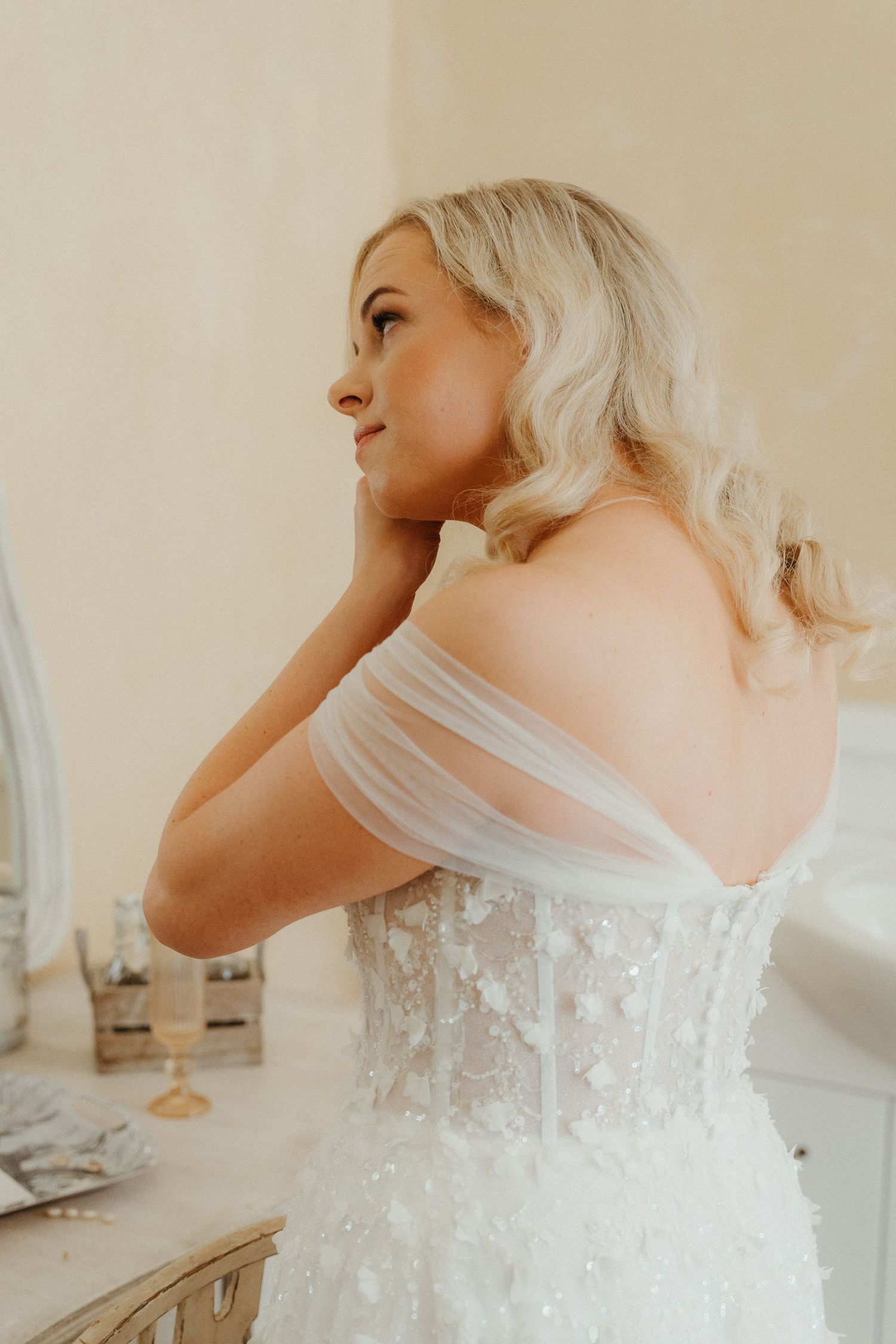Blonde bride in off-shoulder lace wedding gown adjusting earring, viewed from behind in soft natural light.