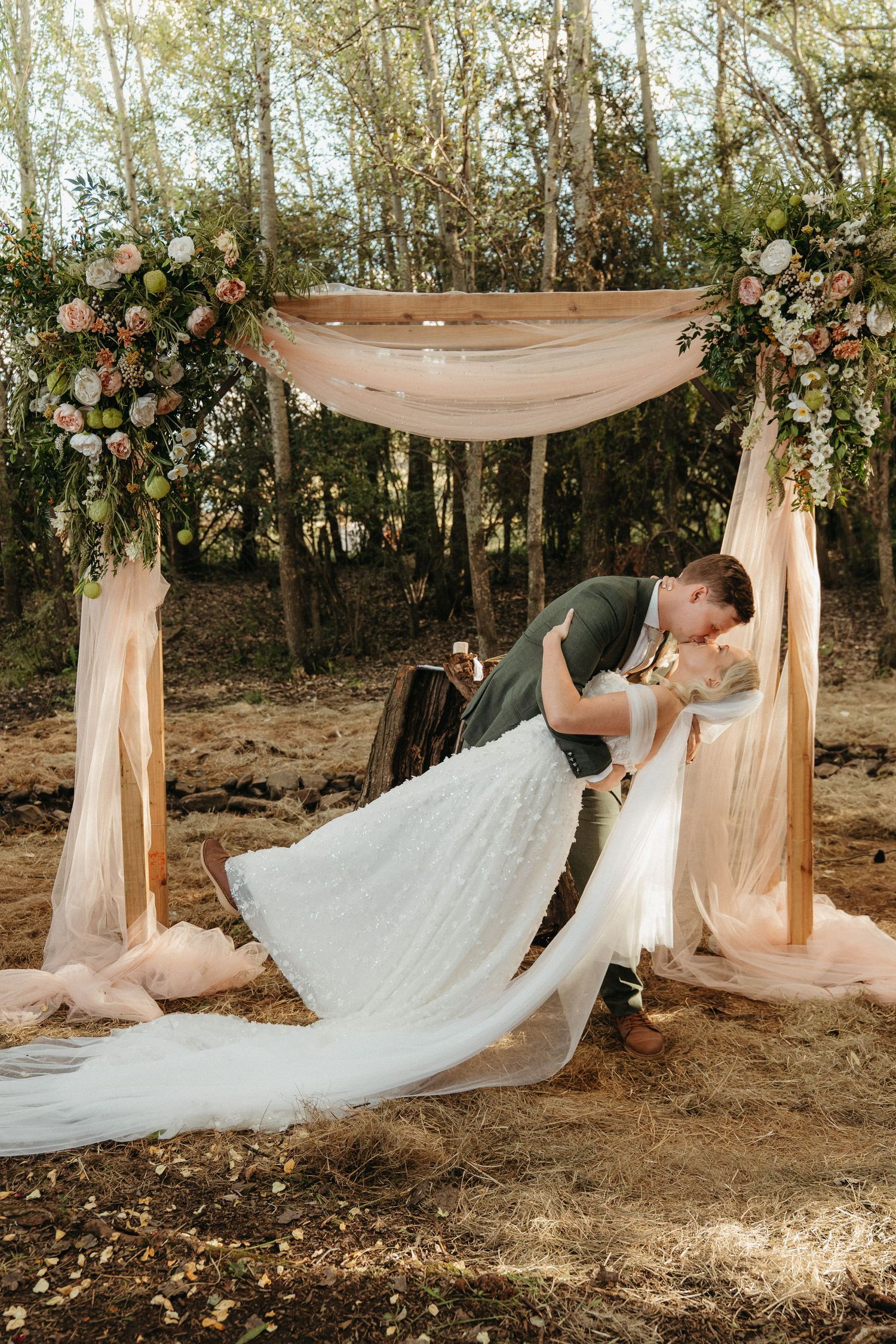 Bride and groom sharing a romantic dip kiss under a floral arch with blush draping in an outdoor woodland wedding ceremony.